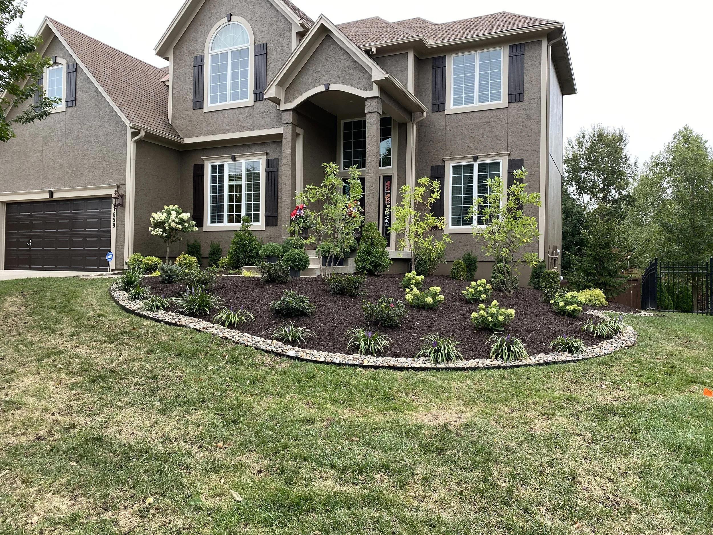 A large, two-story house with a brown shingle roof, gray exterior walls, and white-framed windows. The front yard has well-maintained landscaping with small bushes, flowering plants, and a curved border of gravel. There's a garage on the left side an