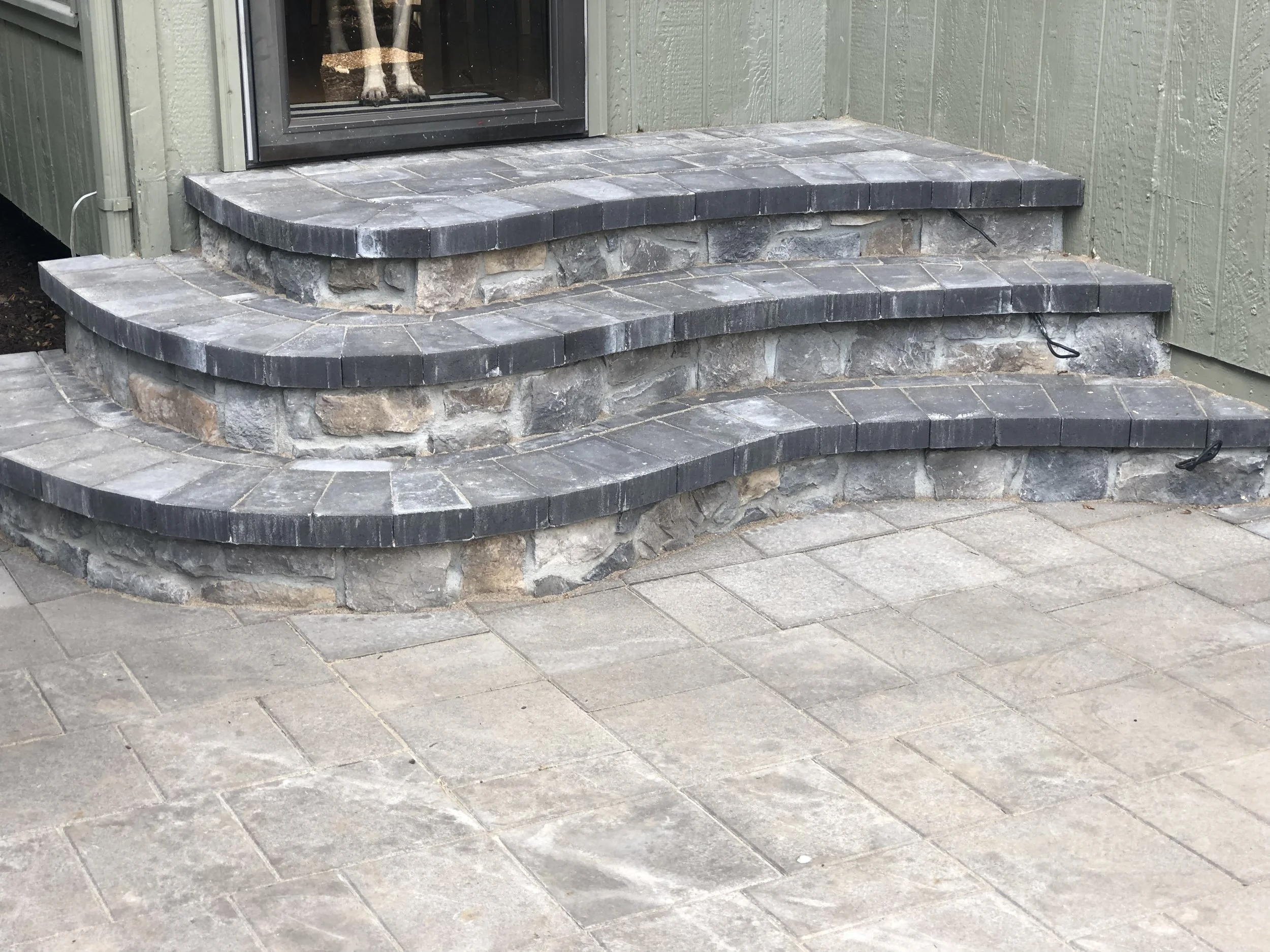 This image shows three stone steps leading to a glass door. The steps are made of dark gray bricks and natural stones, set against light green wooden walls.