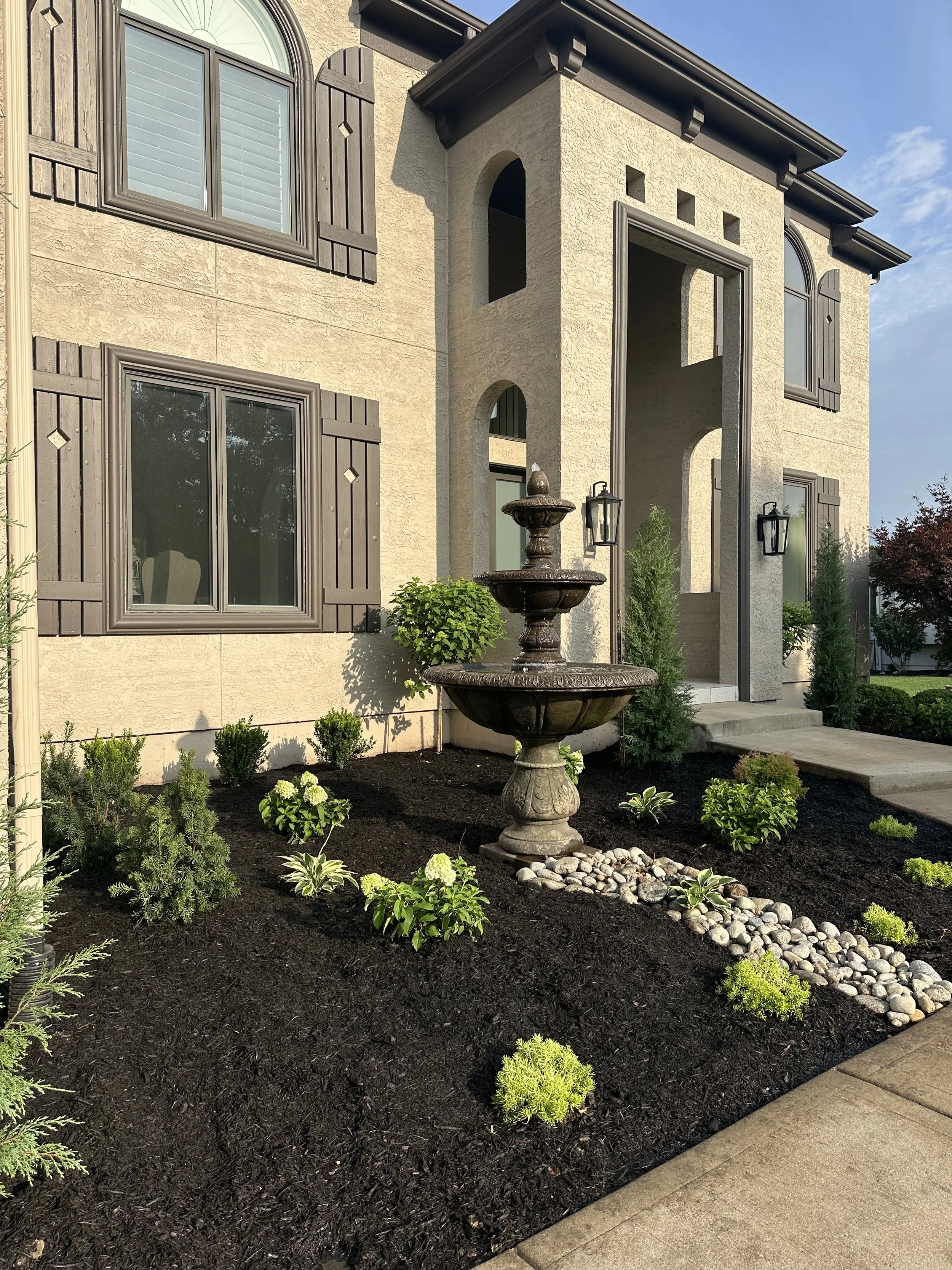 Front yard garden with freshly planted small bushes, decorative rocks, a stone fountain, and a concrete walkway leading to a house with beige textured walls, decorative shutters, and an archway entrance.