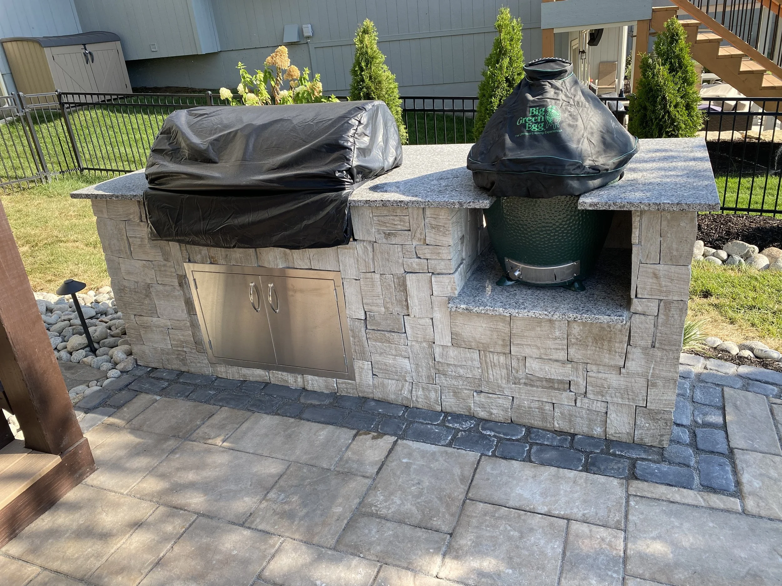 An outdoor kitchen with a stone base, granite countertop, and a black covered grill and smoker. There are small trees and a fenced yard.