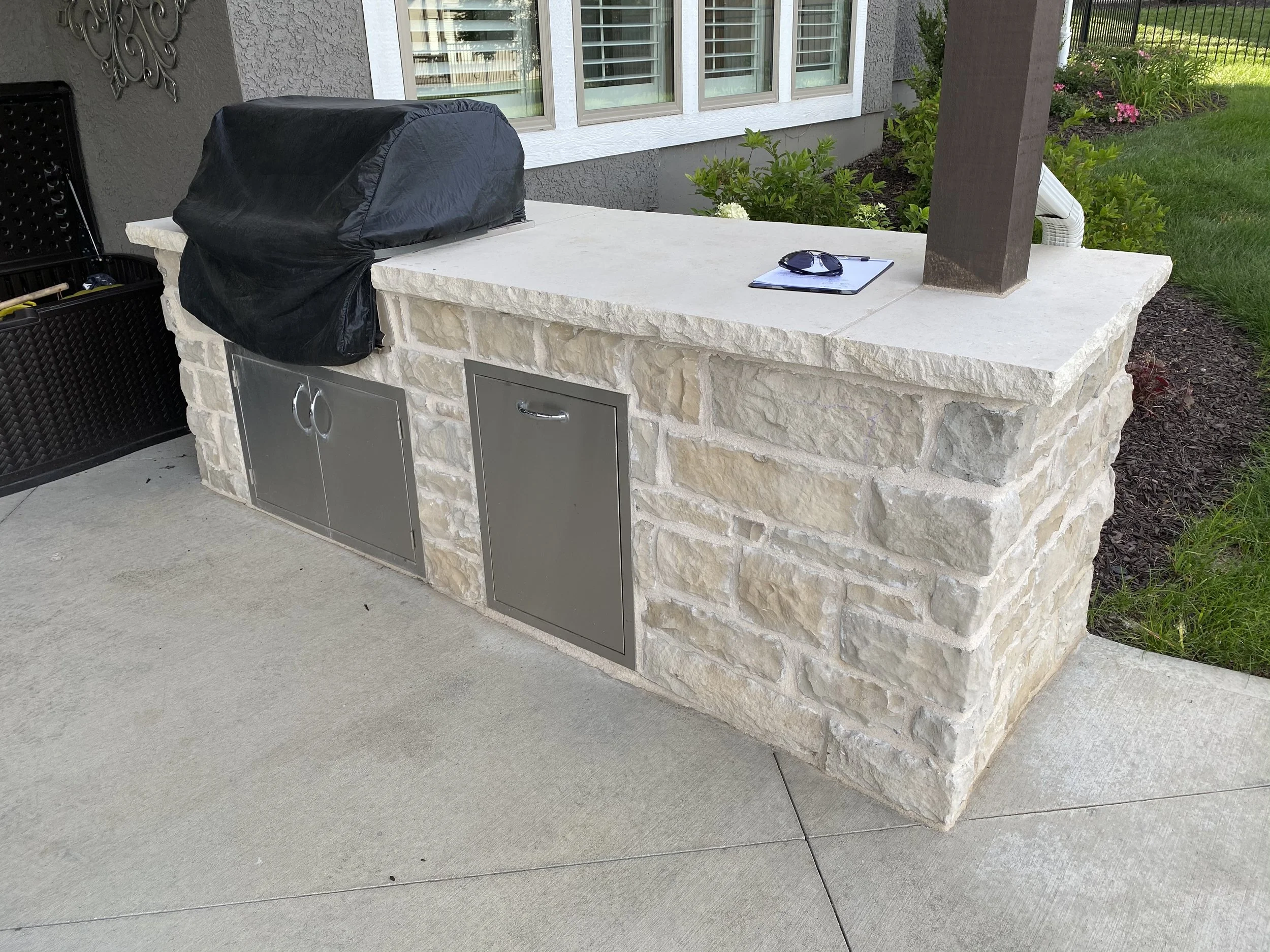 Outdoor kitchen island with a black covered grill, sunglasses, and a clipboard, situated on a concrete patio adjacent to a grassy yard with plants and flowers.