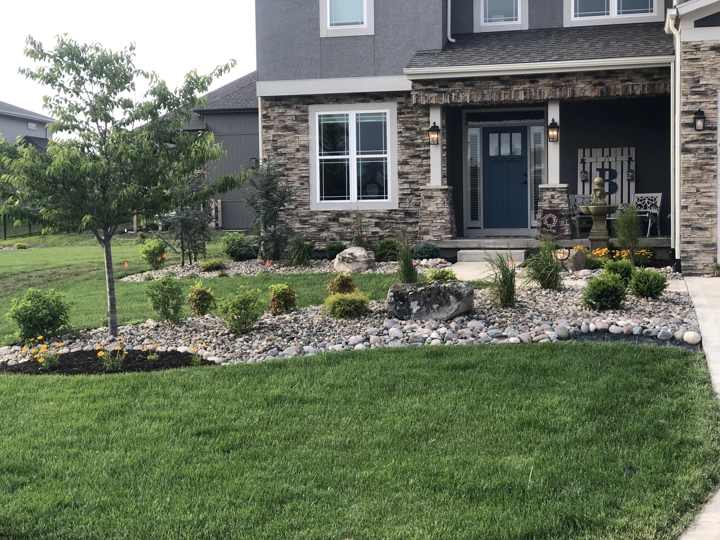 Front yard of a modern house with a lawn, small trees, and landscaping stones, with the house's exterior featuring stone and gray siding.