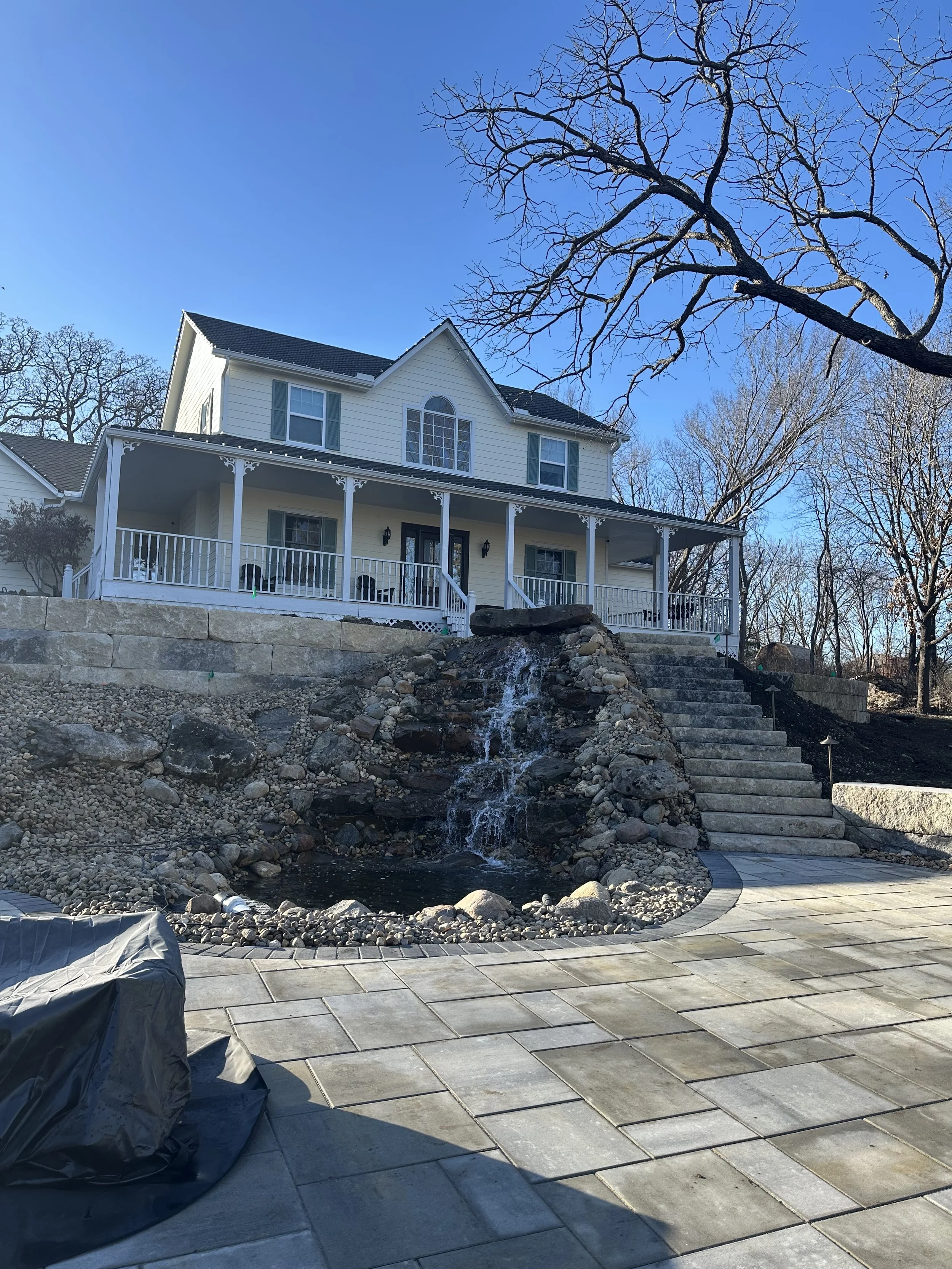View of a house with a large front porch, white siding, and dark shutters. In front, there is a landscaped area with rocks, a small waterfall, and a paved patio. A leafless tree extends across the top right of the image, and the sky is clear and blue