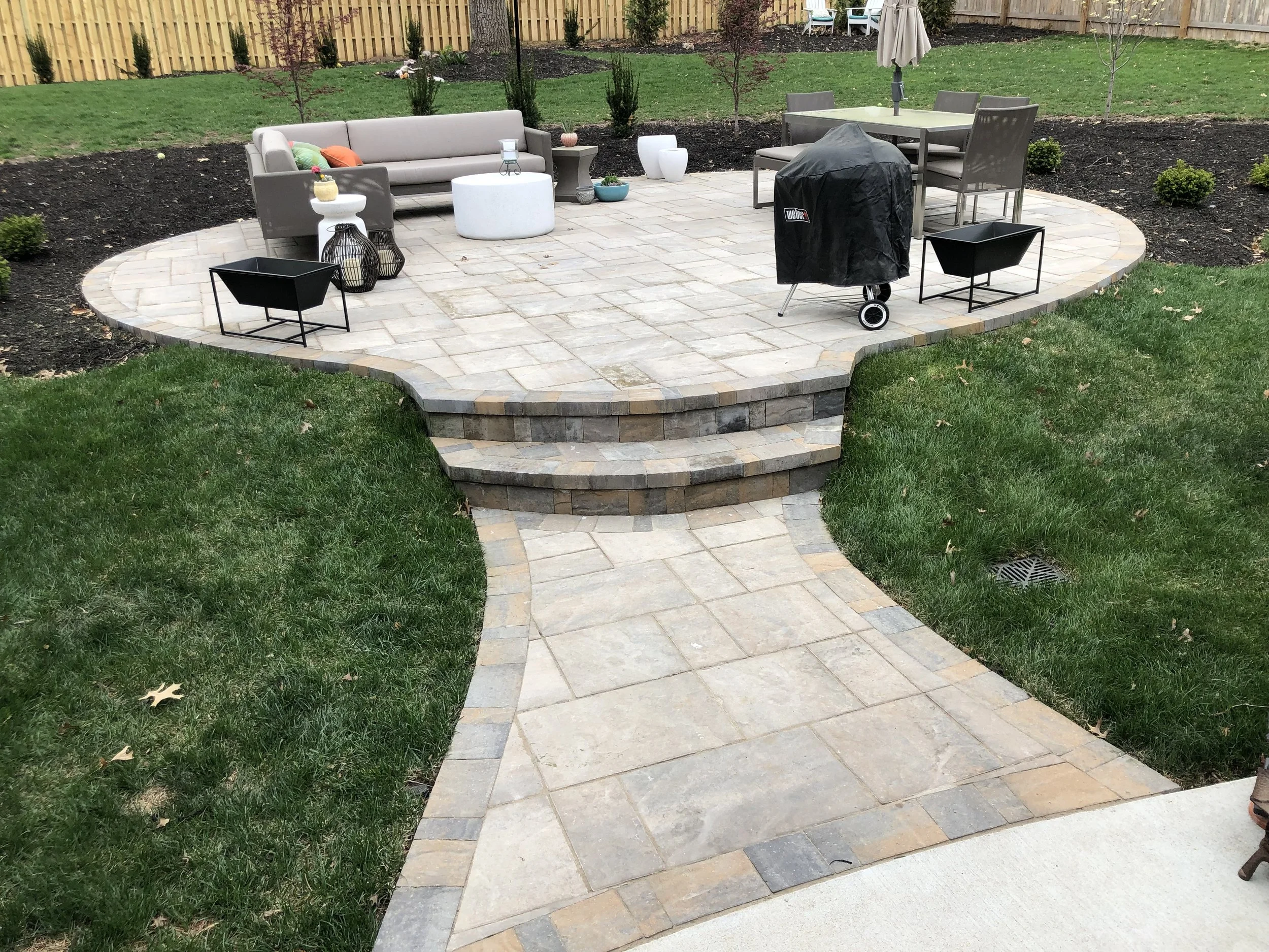 A backyard patio with a light grey sofa, small coffee tables, decorative lanterns, and a black grill covered with a black cover, surrounded by a garden with trees, shrubs, and a wooden fence.
