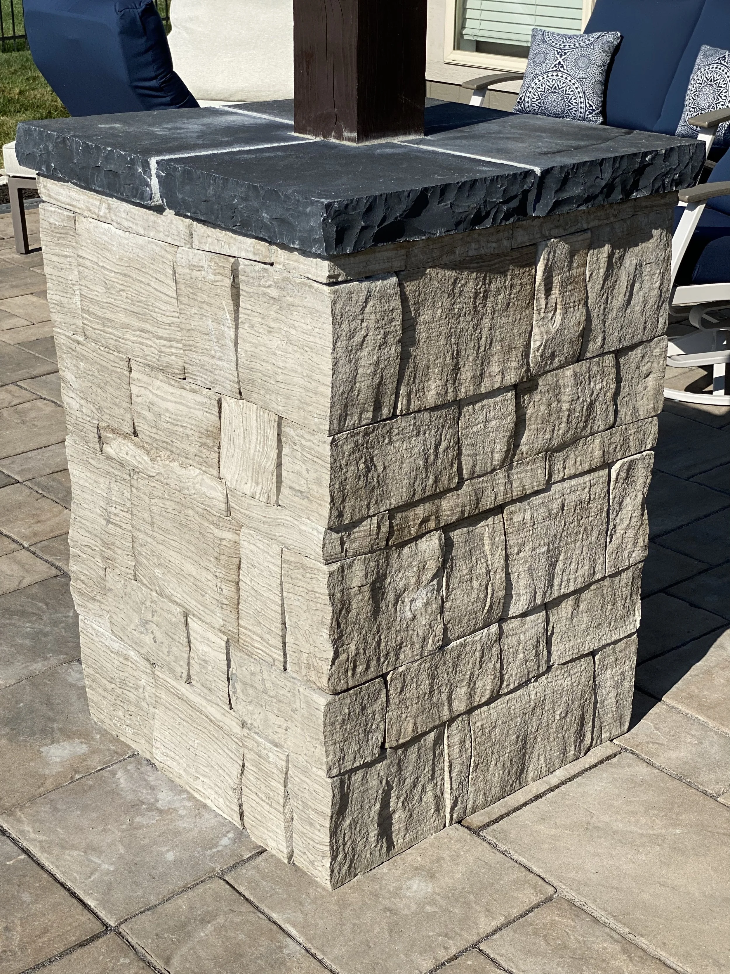 A stone outdoor table with a dark gray stone top and a base made of beige, textured stone blocks, situated on a paved patio area.
