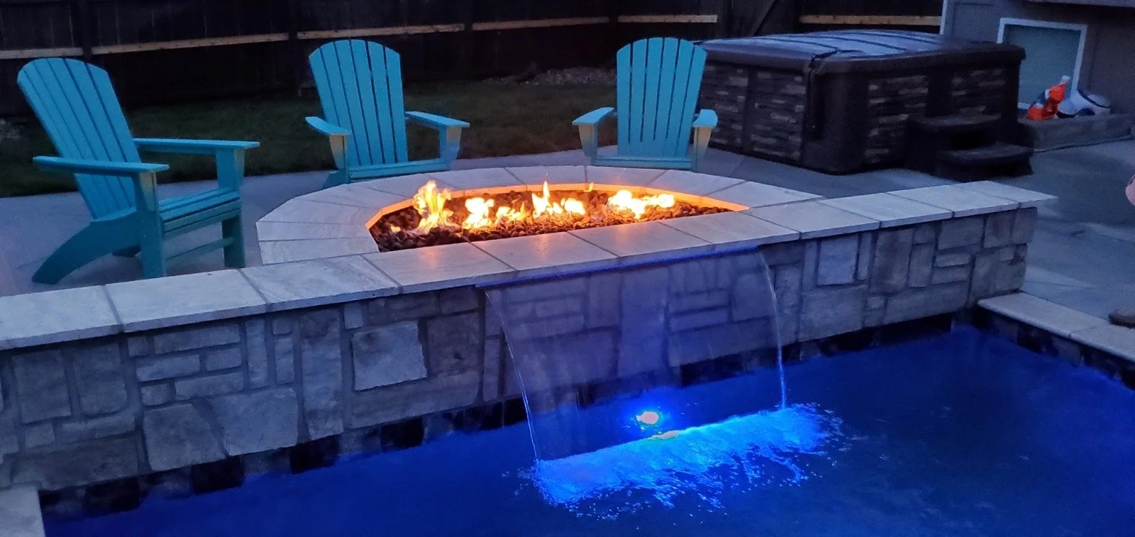Outdoor patio with stone fire pit, three light blue Adirondack chairs, water feature with falling water, and a hot tub in the background at twilight.