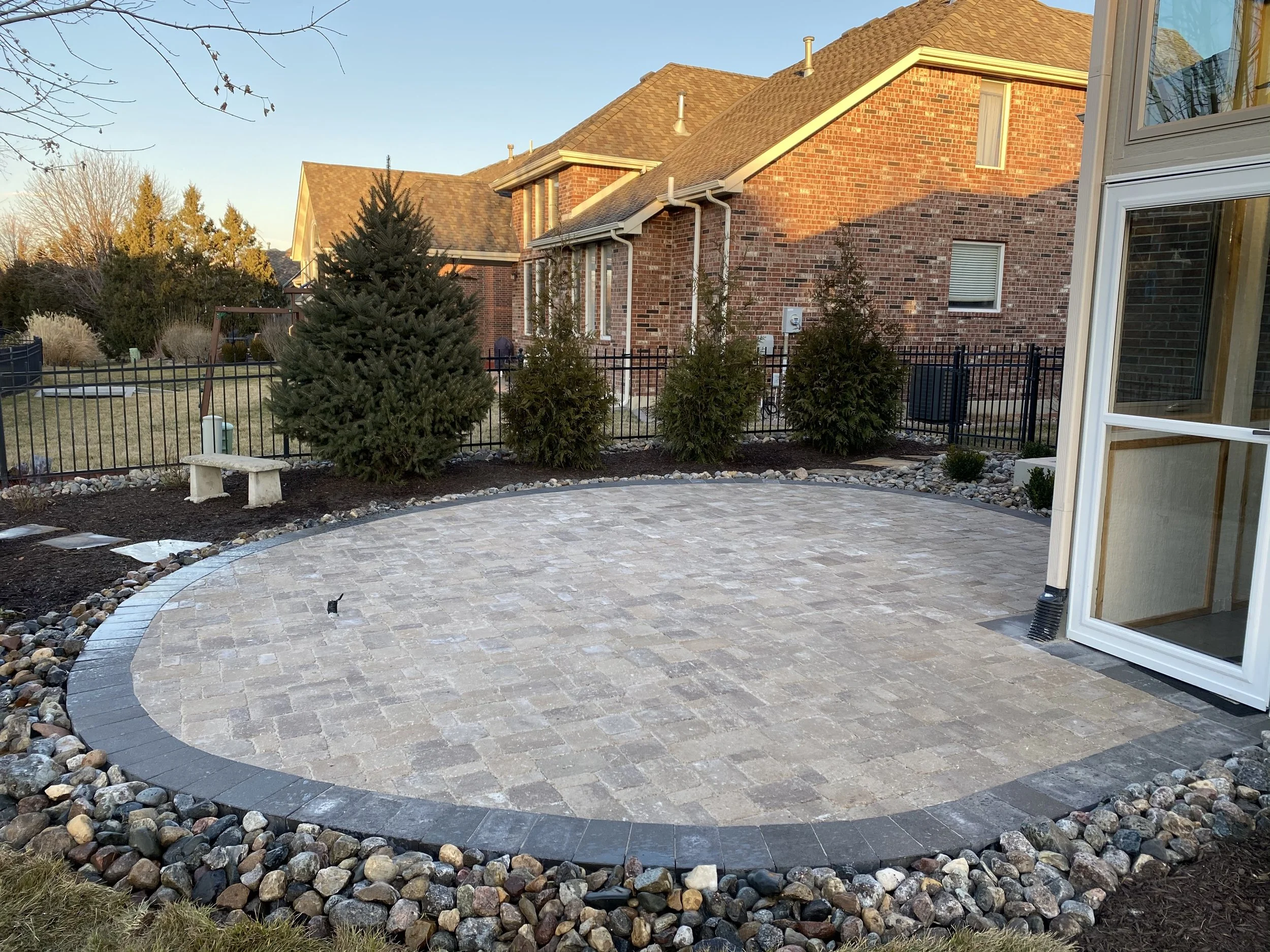 Backyard with a paved patio, landscaped garden, evergreen trees, and a brick house during sunset.
