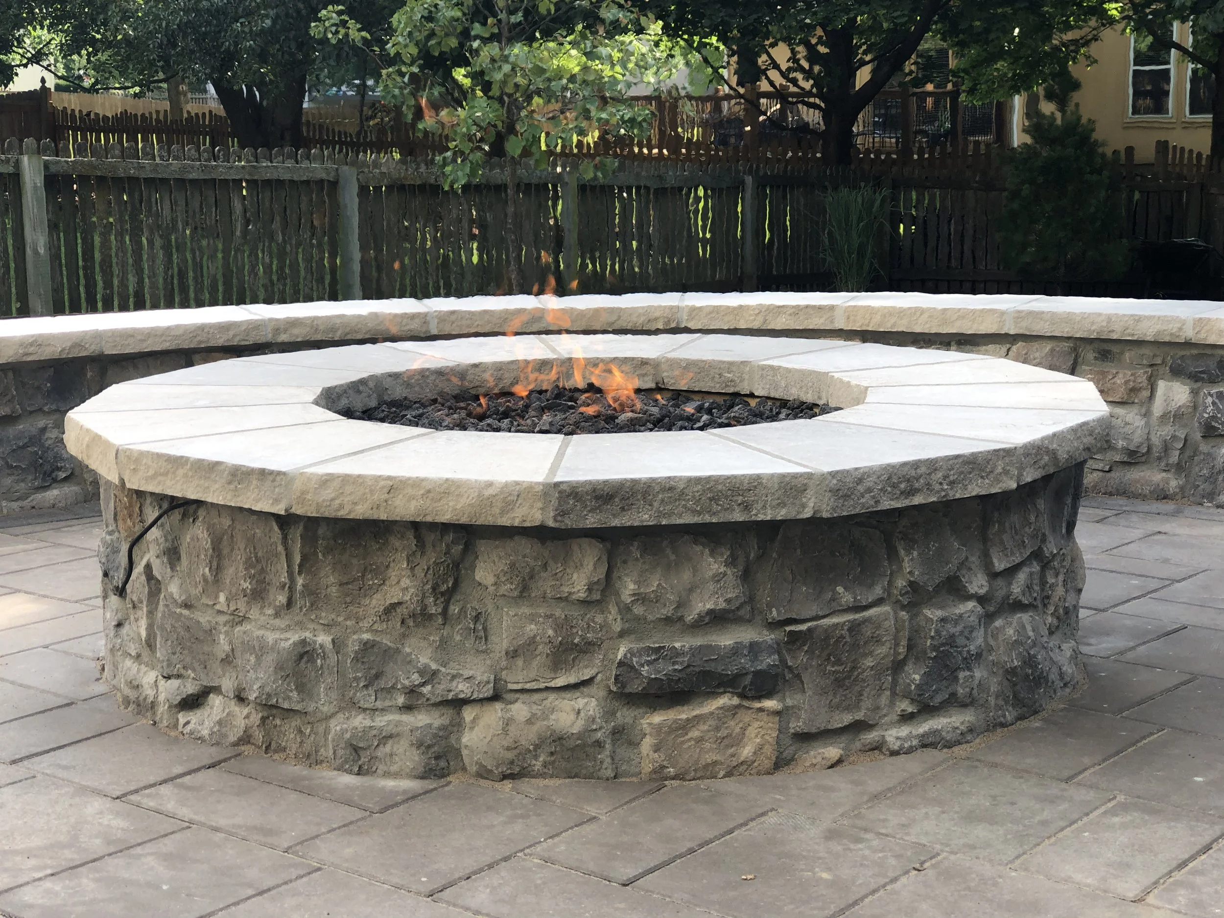 A stone fire pit with flames, situated on a patio with wooden planks. In the background, there are trees, a wooden fence, and a house with windows.