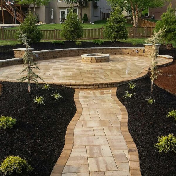 A landscaped backyard with a paved walkway leading to a circular paved area with a built-in fire pit. The area is surrounded by small trees and mulched garden beds, with a wooden fence and neighboring houses in the background.