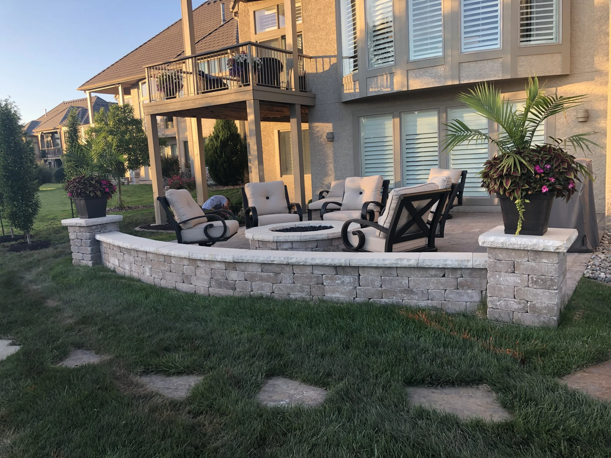 A backyard patio area with cream-colored cushioned chairs arranged around a fire pit, surrounded by a curved stone wall. Large potted plants with colorful flowers are positioned at the corners of the patio, which is adjacent to a beige house with whi