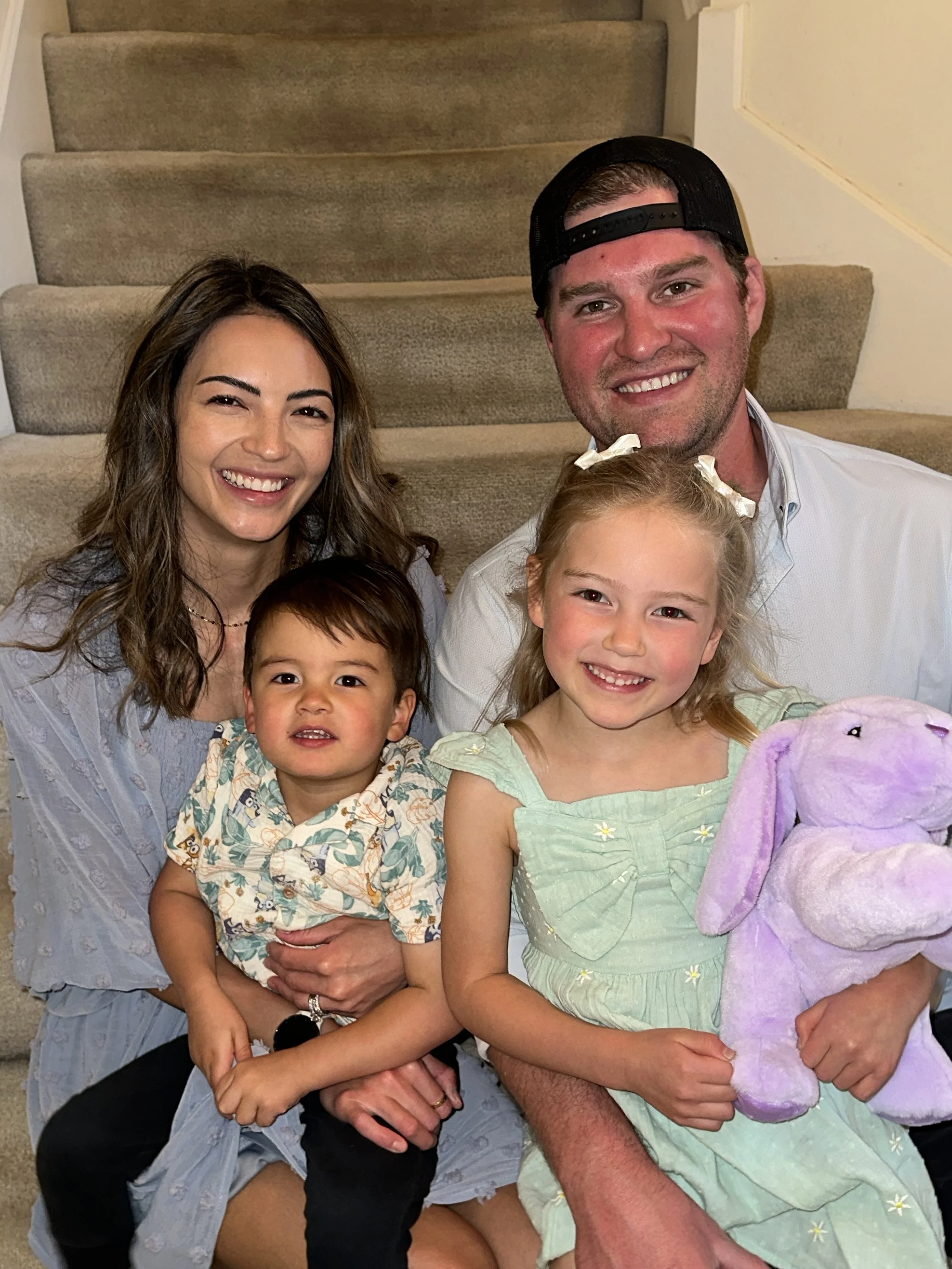A group photo of two children and two adults sitting on carpeted stairs, smiling, with the children holding plush toys; a little girl on the right holding a purple stuffed animal, and a young boy on the left being held by a woman with long brown hair, wearing a patterned shirt. The man on the right is wearing a backward black cap and a white shirt, with his arm around the girl.
