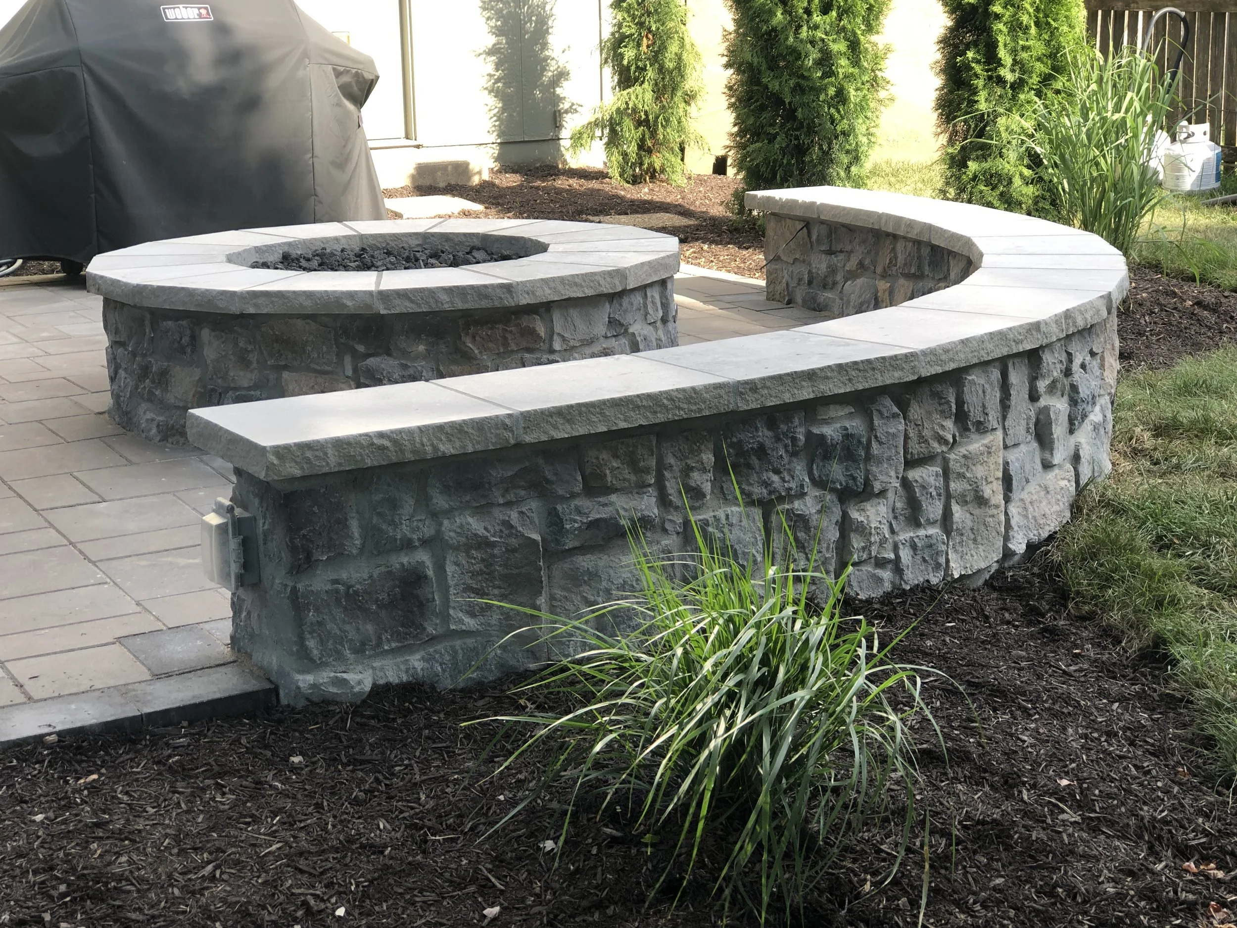 Outdoor fire pit with a stone base and a stone ledge, surrounded by mulch, grass, and shrubs, with a covered barbecue grill nearby.