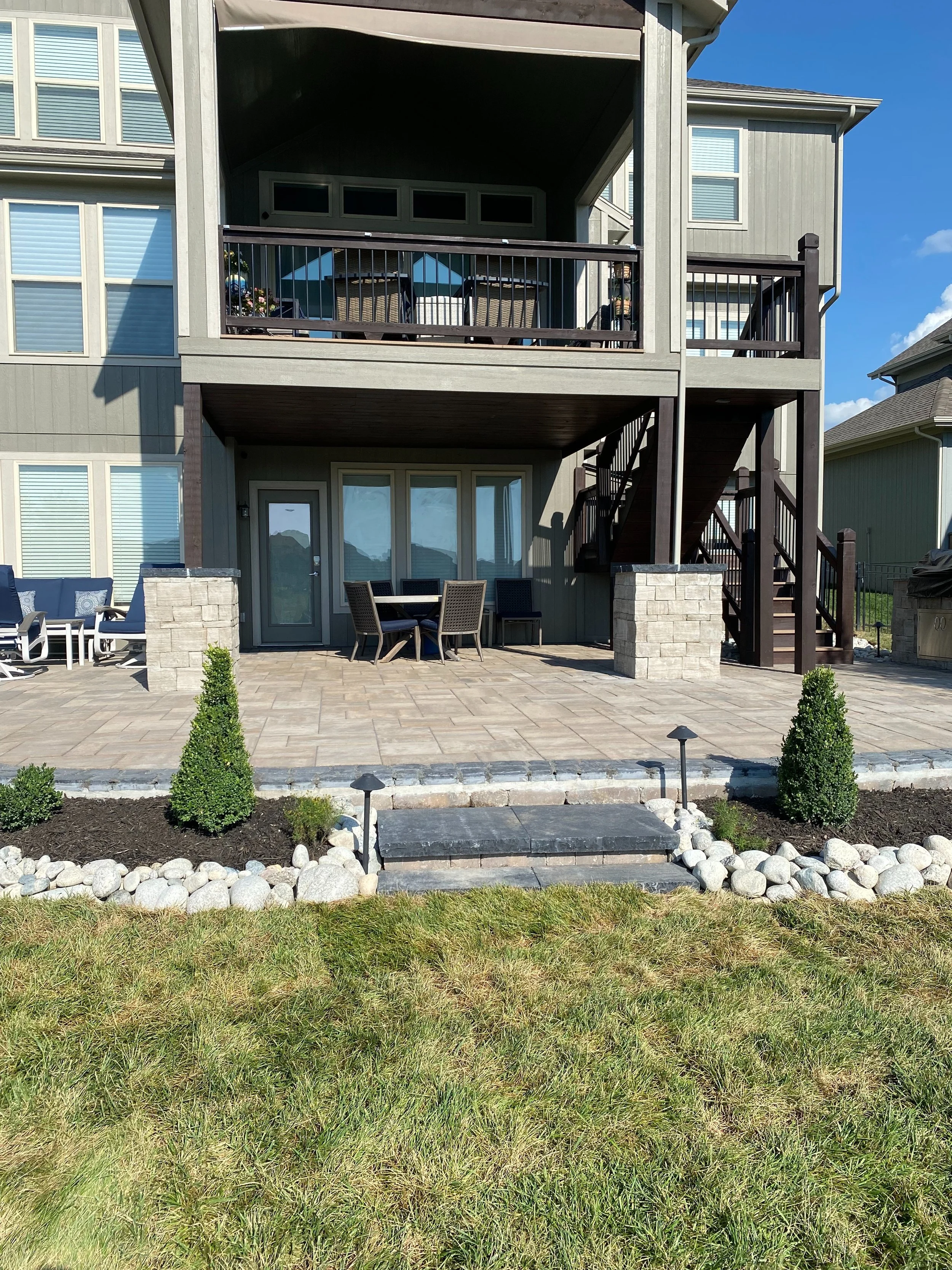 The image shows the backyard of a modern house with a two-story patio. The patio has a sitting area with outdoor furniture, including a table with chairs and a blue umbrella, and a dining table with chairs. There is a staircase on the right side lead