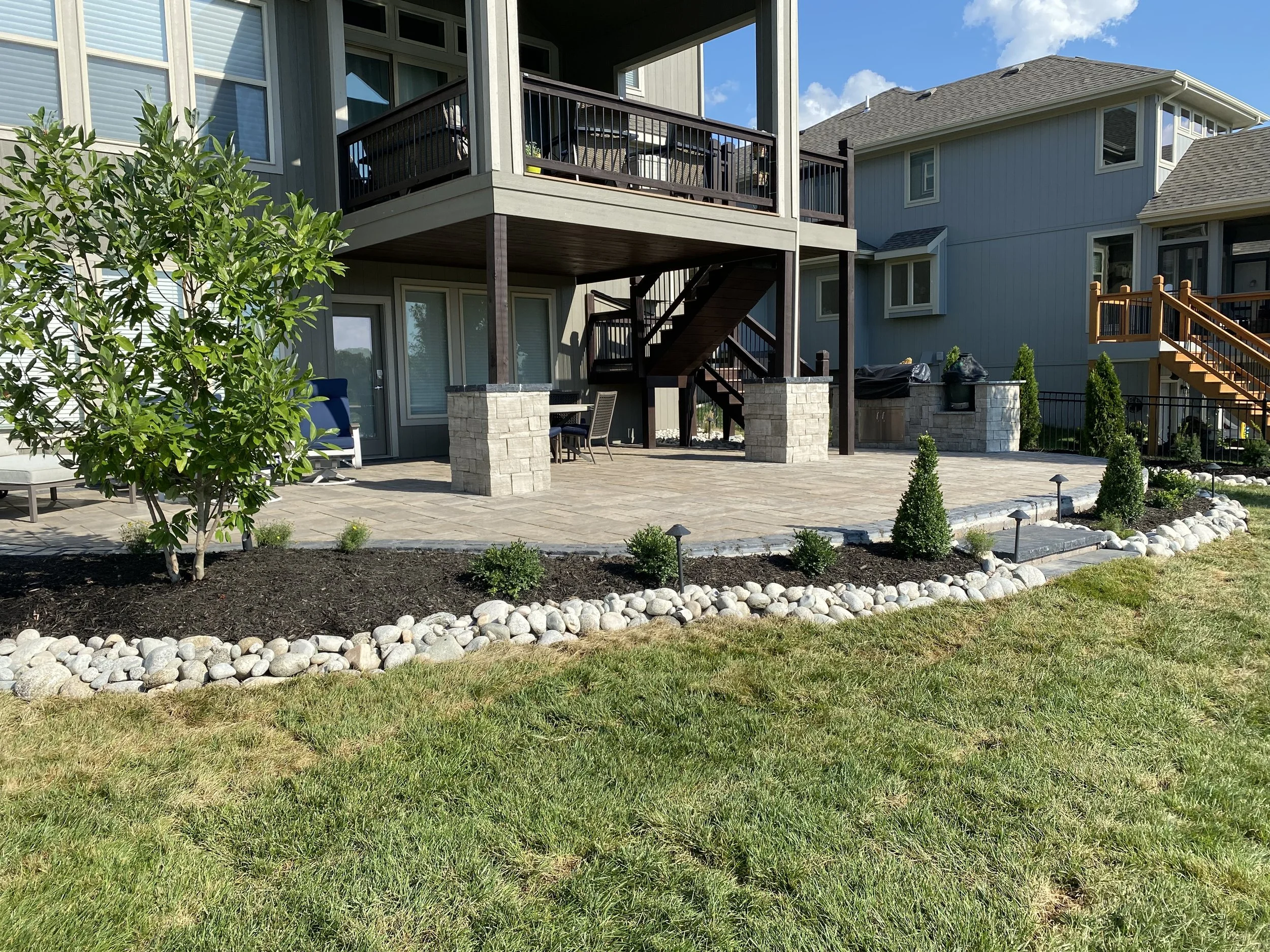 A backyard patio with a paved stone floor and a landscaped garden border with small shrubs and rocks. There is a modern gray house with large windows and a covered deck with stairs leading down to the patio, and grill equipment.