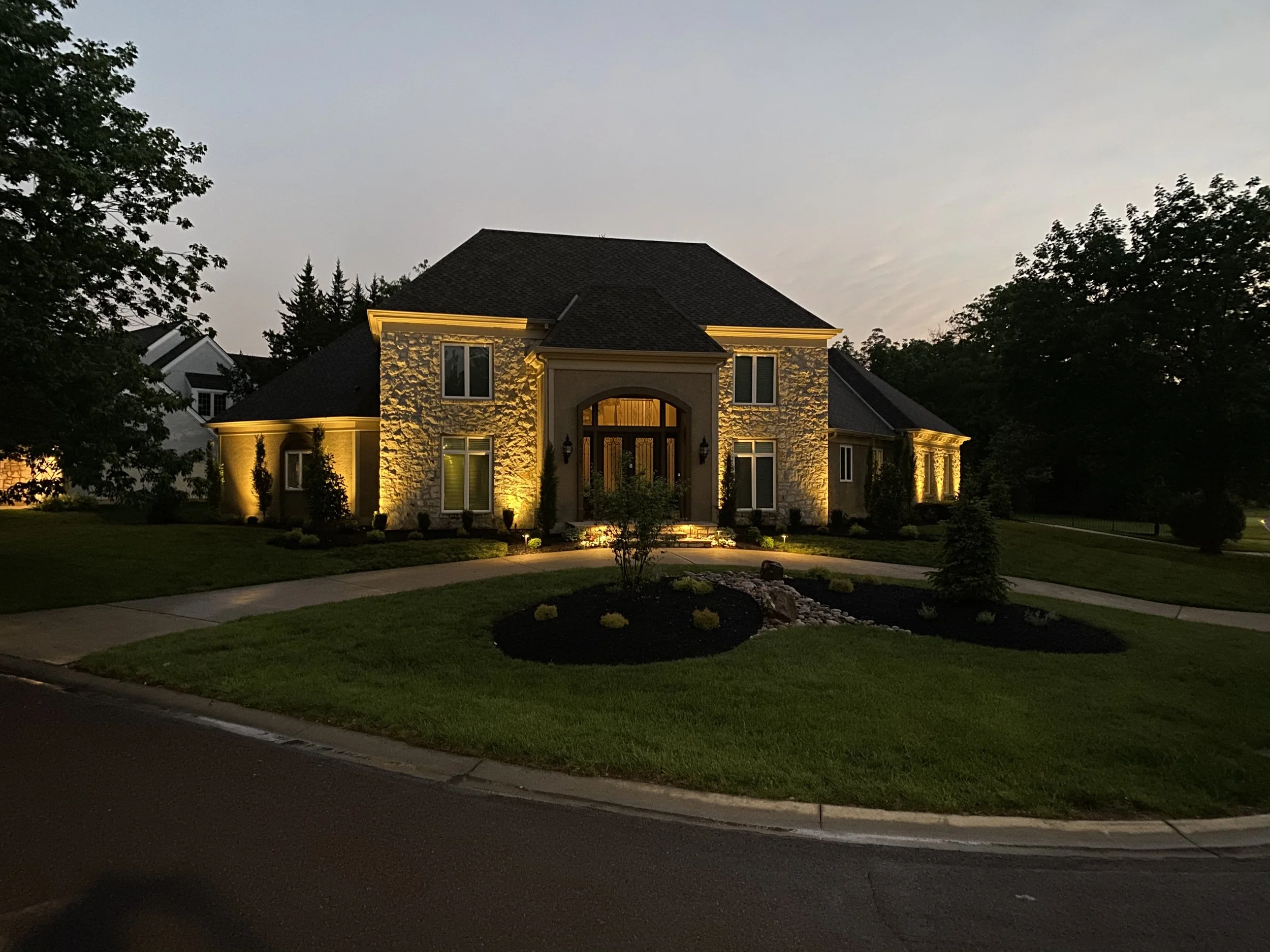 A house with exterior lighting illuminating the stone facade and driveway at dusk, surrounded by trees and a well-manicured lawn.