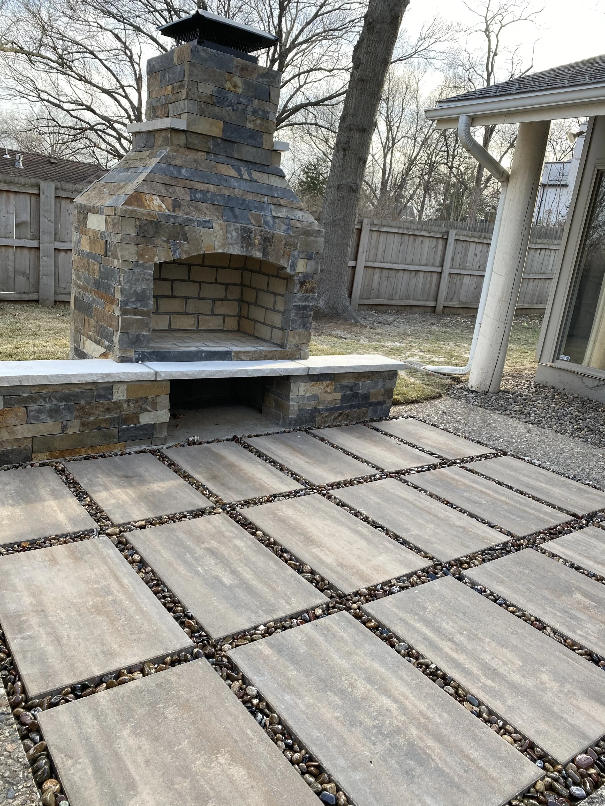 An outdoor fireplace with a brick and stone chimney, a stone hearth, and a stone pathway leading to it in a backyard, with a wooden fence and leafless trees in the background.