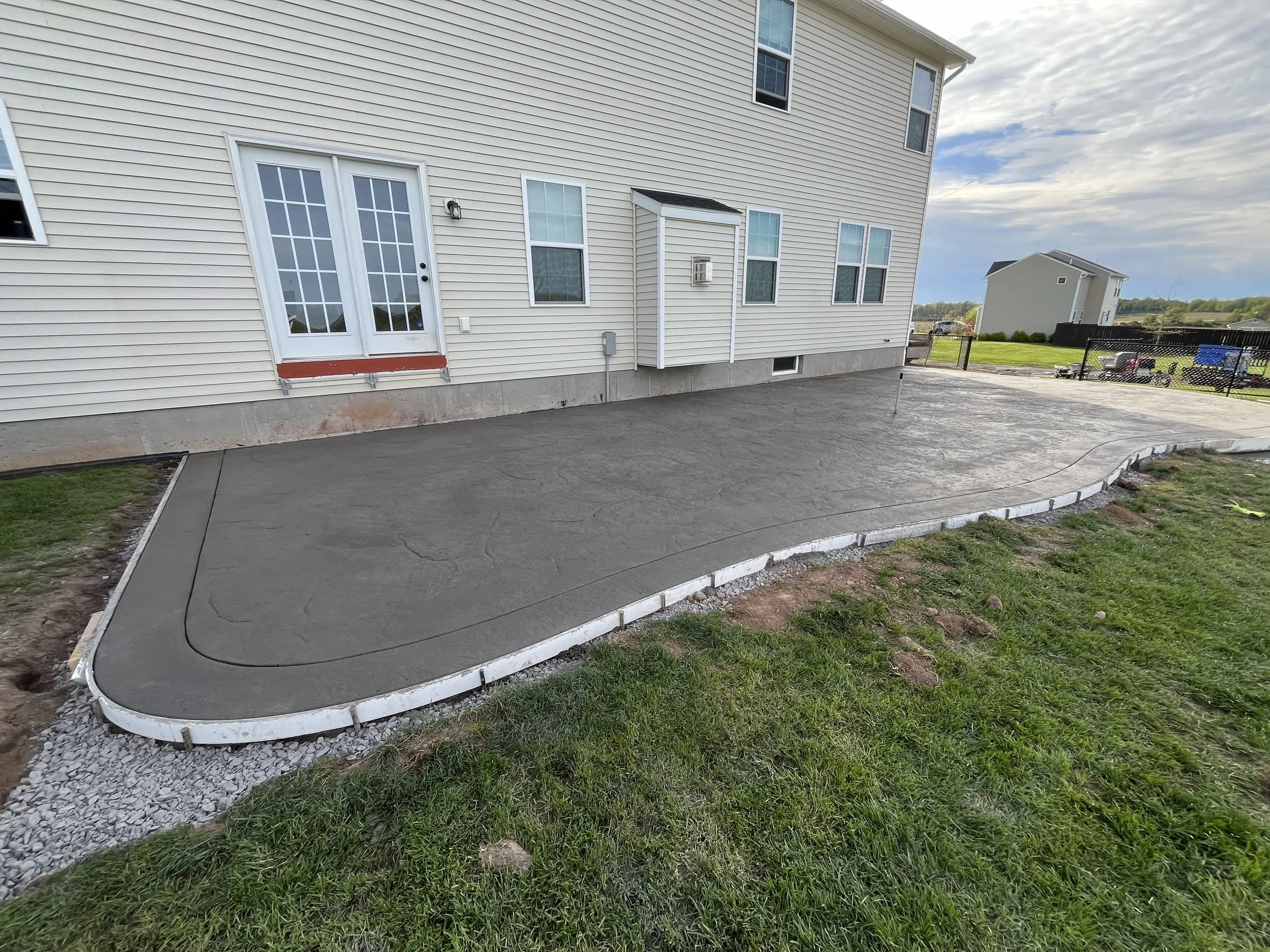 Newly poured concrete patio in backyard with curved edges, adjacent to a white house with sliding glass doors and windows, and some grass and dirt around the edges.