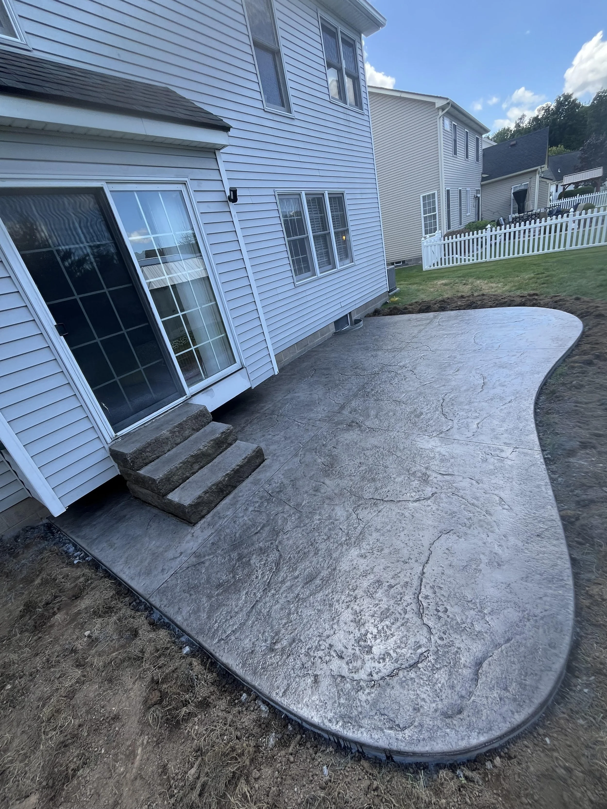 Freshly poured concrete patio with curved edges outside a two-story house, with stairs leading to sliding glass doors. The house has white siding and multiple windows, and other similar houses are visible in the background.
