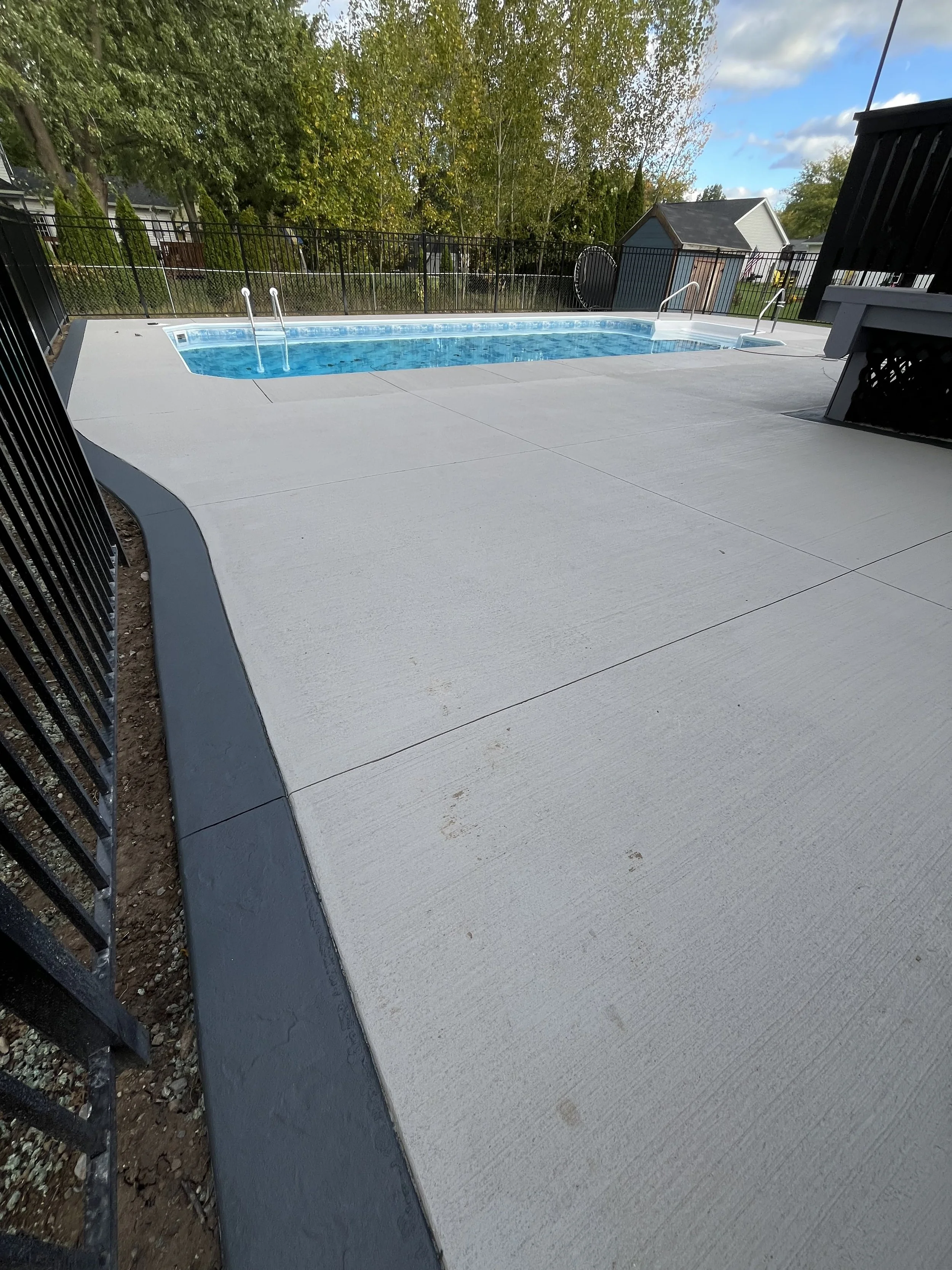 Personal backyard swimming pool with concrete deck, surrounded by a black metal fence, green trees, a small shed, and neighboring houses in the background under a partly cloudy sky.