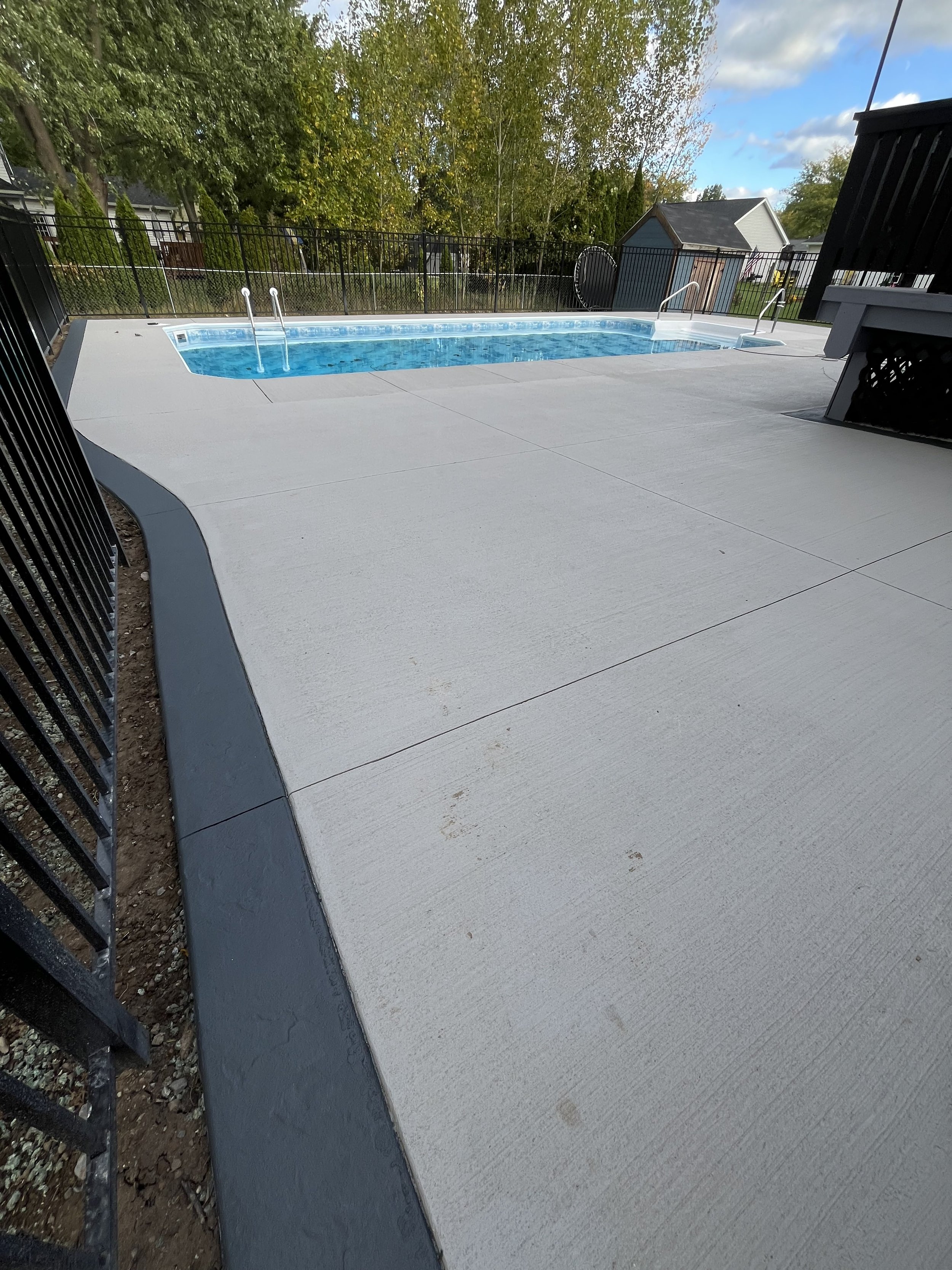 Empty backyard pool with a concrete deck and black metal fence, surrounded by trees and a small shed in the background.