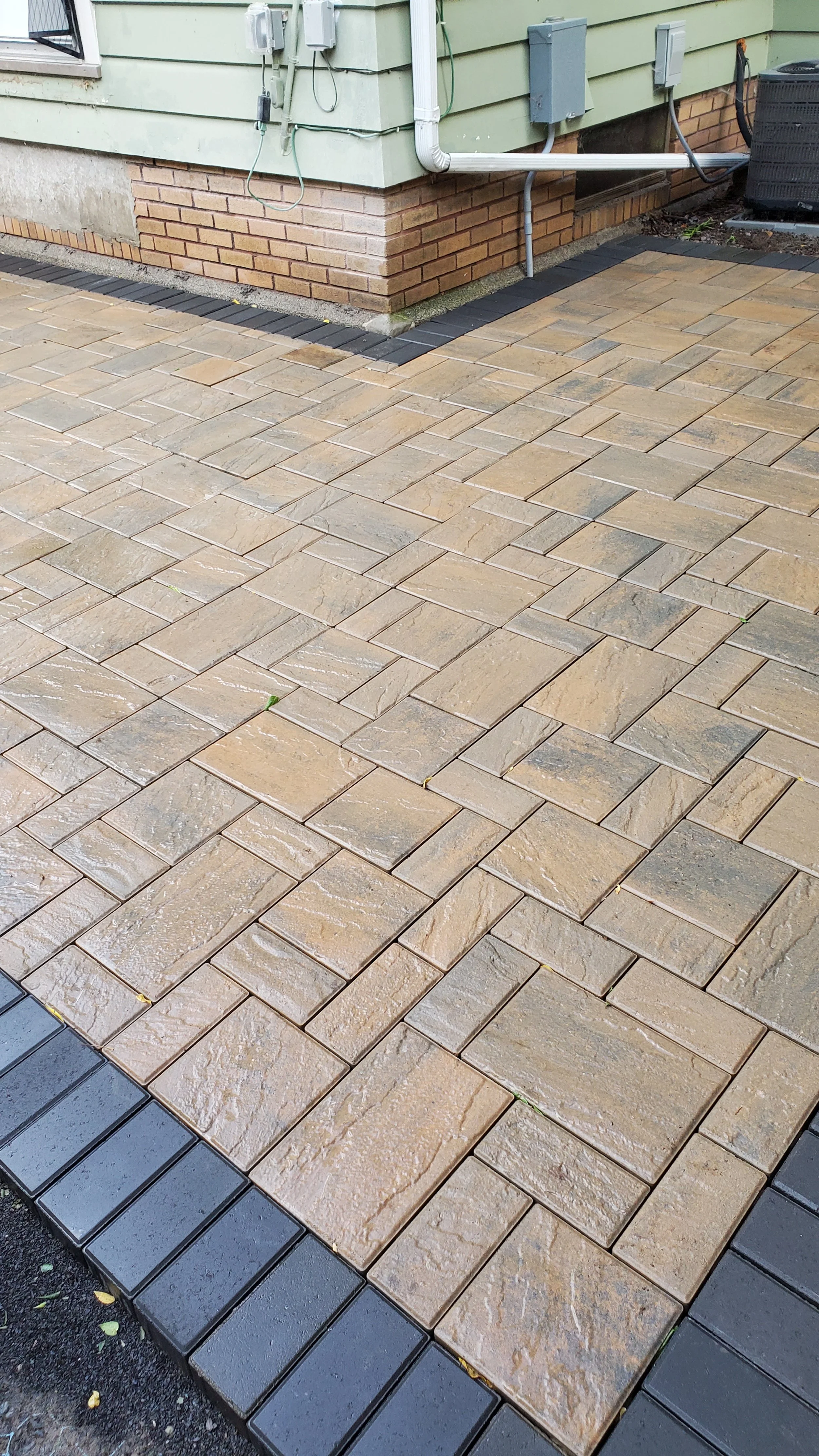 Patio with tan and black stone pavers arranged in a pattern next to a house with green siding and brick foundation, featuring an air conditioning unit and utility boxes.