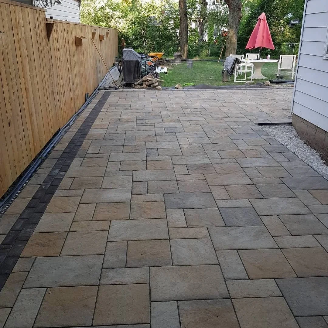 Patterned stone patio in backyard with wooden fence, grass area, outdoor furniture, and trees in the background.
