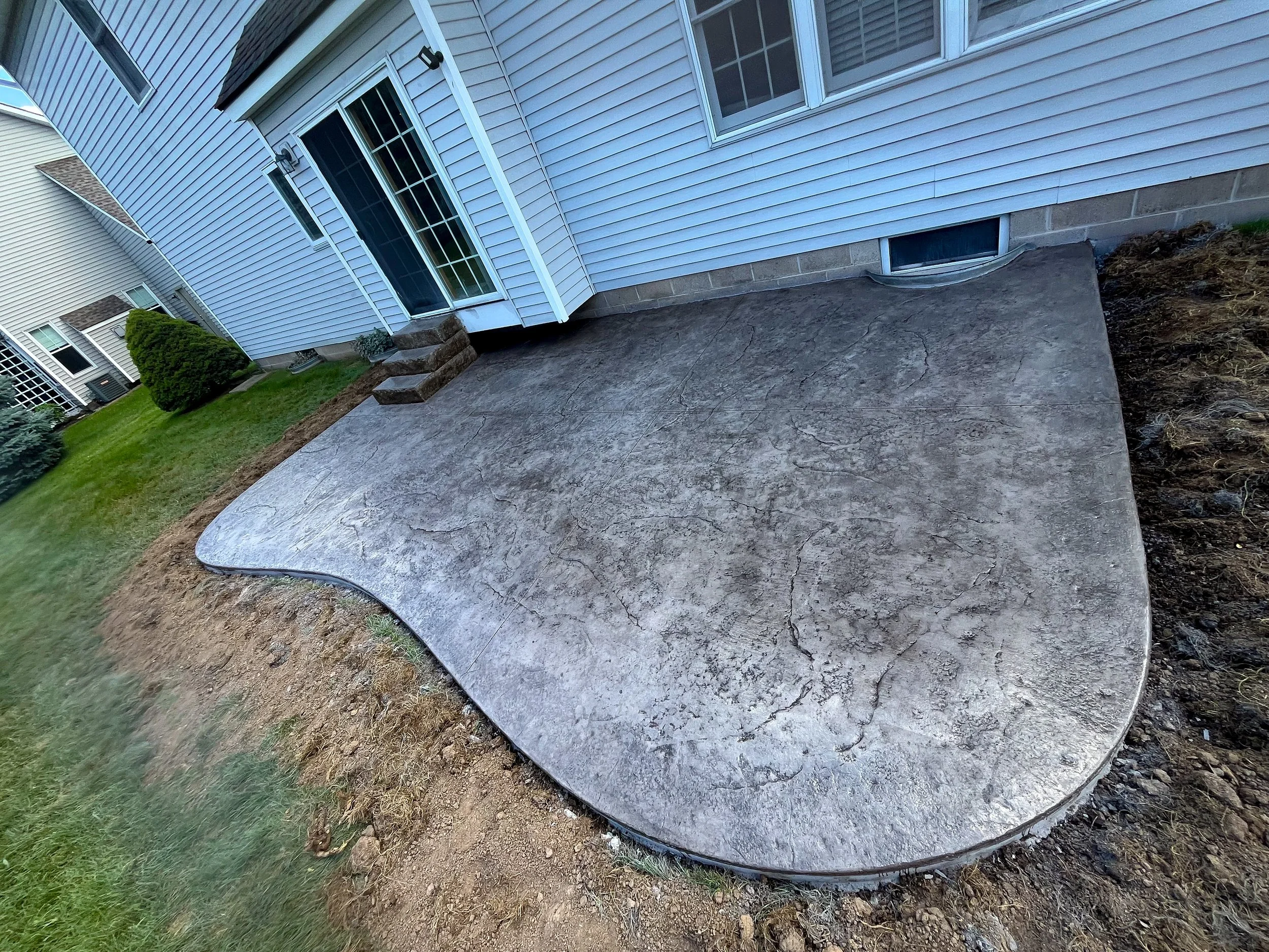 Freshly poured concrete patio in the backyard of a house, with steps leading to a sliding glass door.