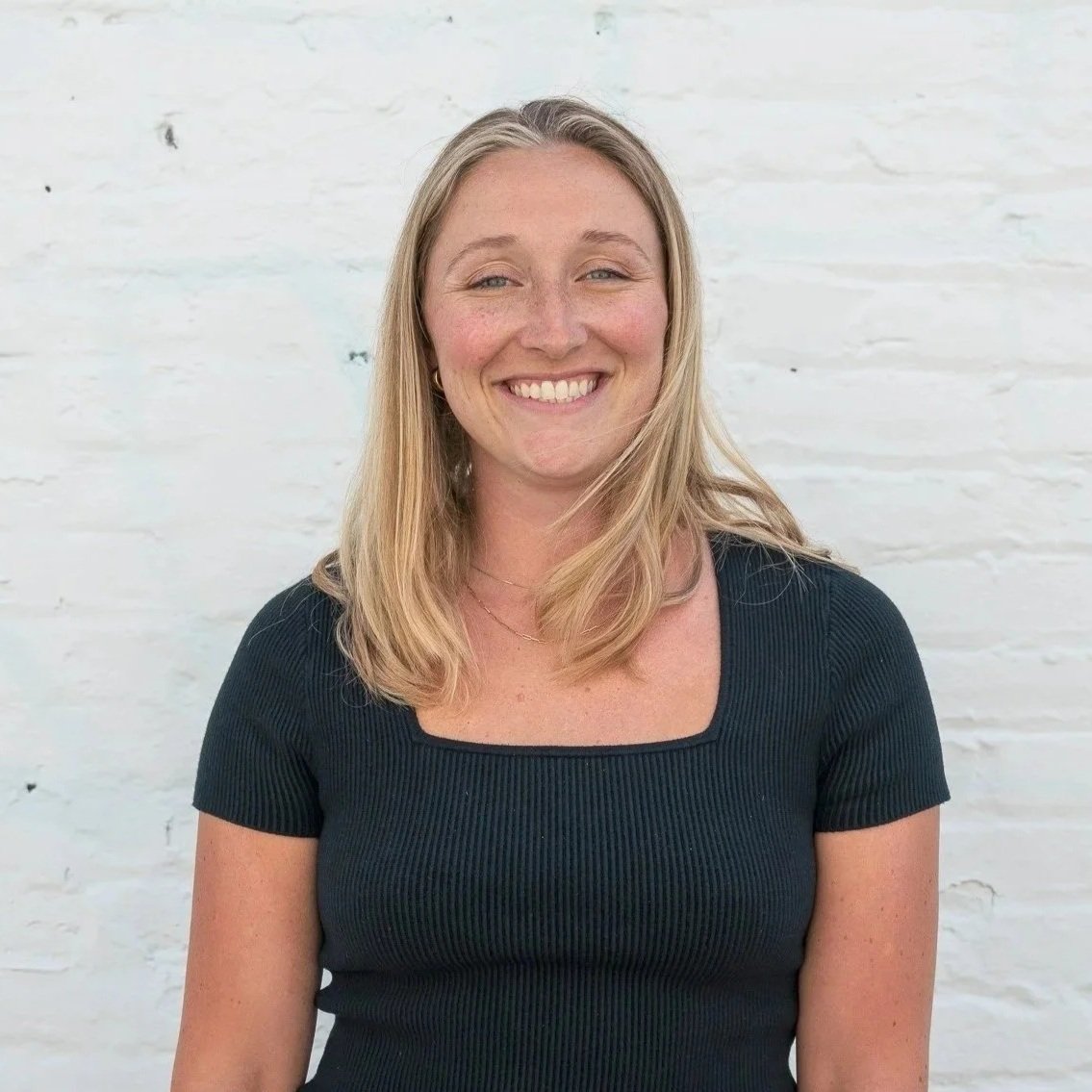 A smiling woman with blonde hair wearing a black top, standing against a white brick wall background.