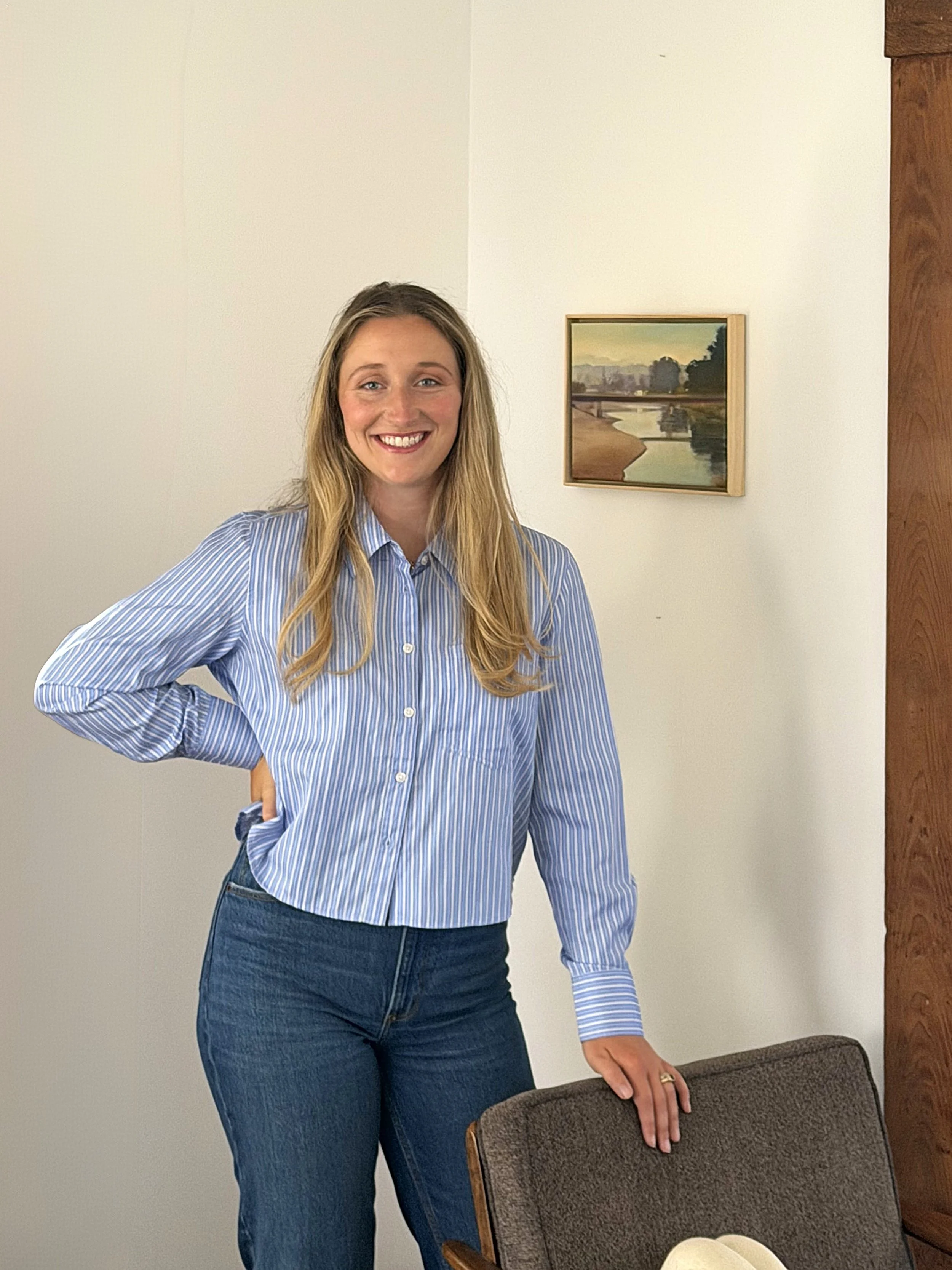 A woman with long blonde hair, wearing a blue and white striped shirt and blue jeans, smiling and standing next to a dark brown chair with her hand resting on its back.