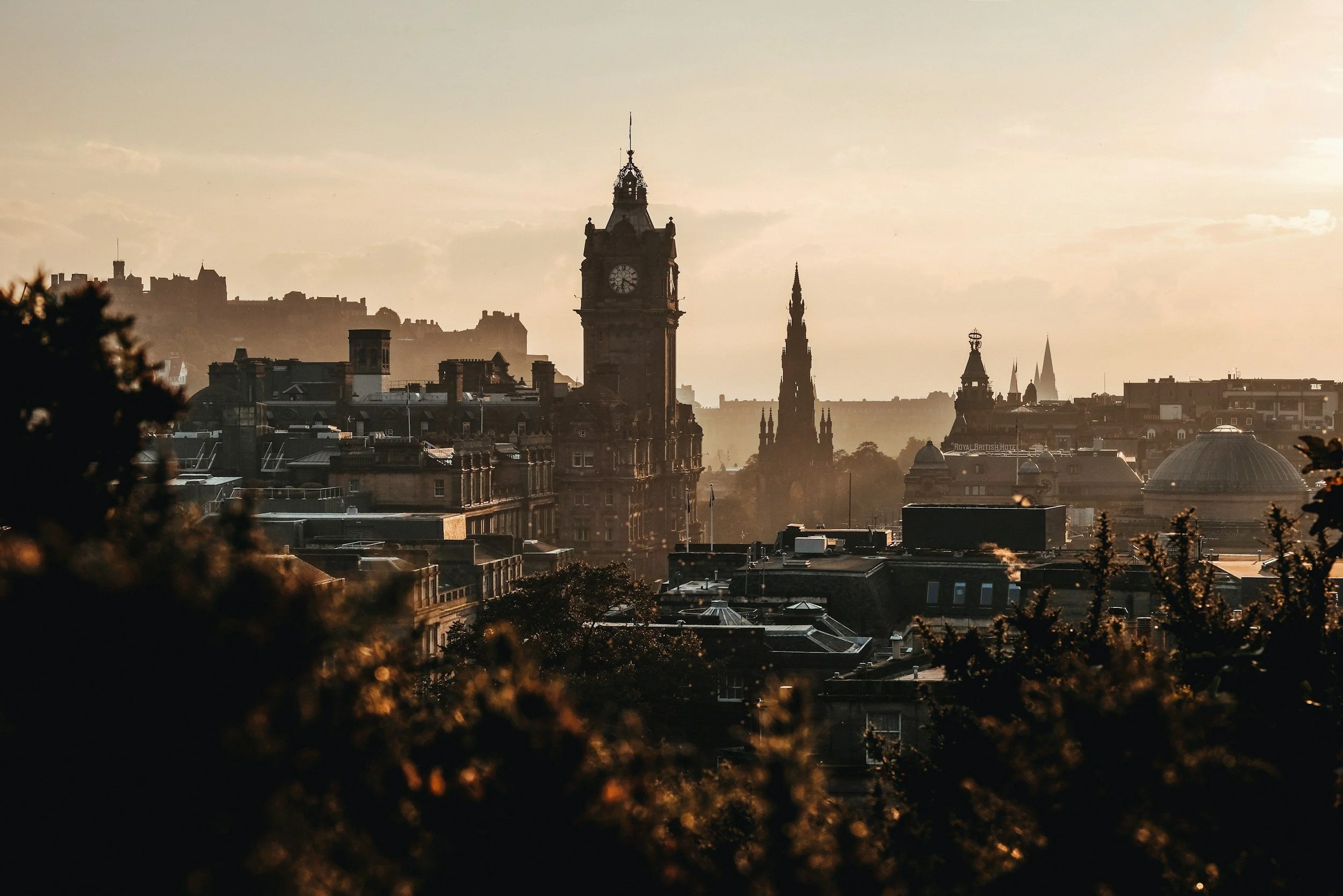 City skyline at sunset featuring historic buildings, clock tower, and spires, with trees in the foreground.