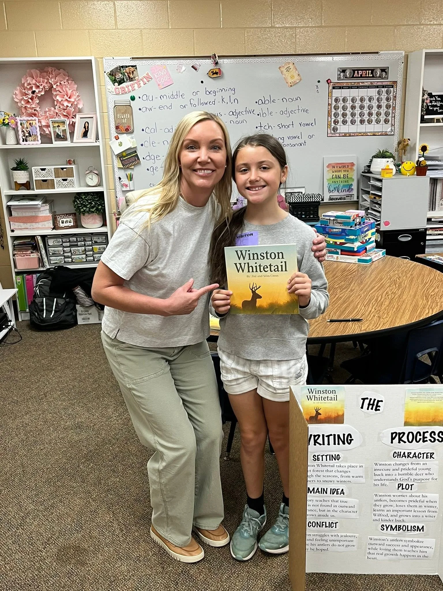 A young girl and an adult woman standing in a classroom, smiling for a photo. The girl holds a book titled 'Winston Whitetail' by Zoe and Silas Cross. In the background, a whiteboard displays notes about the grammar topic, and there are shelves with plants, picture frames, and school supplies.