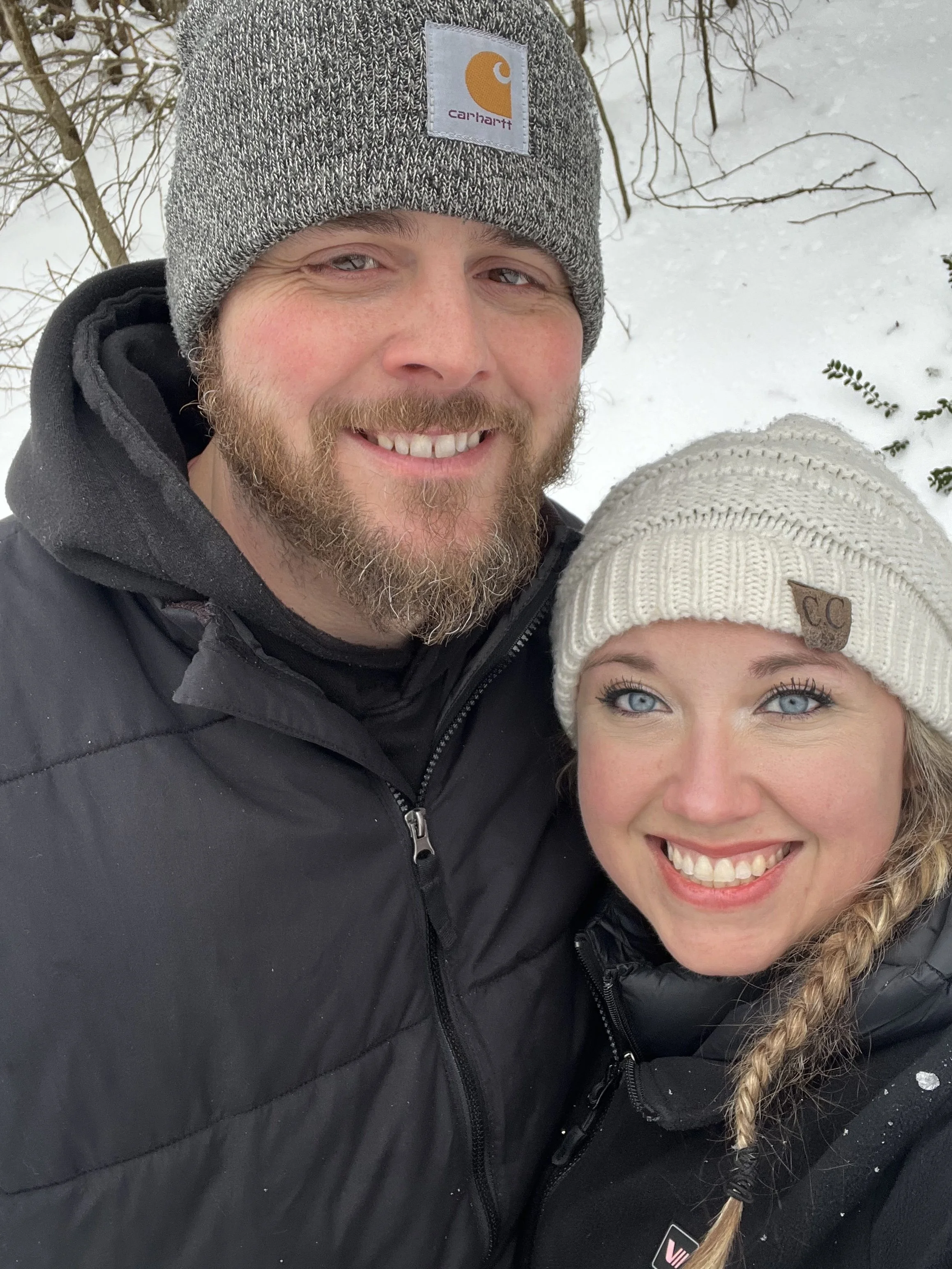 Close-up of a smiling man and woman outdoors in a snowy environment, both wearing beanies and winter jacket. The man's beanie has a Carhartt logo, and the woman's beanie has a small leather patch with initials.