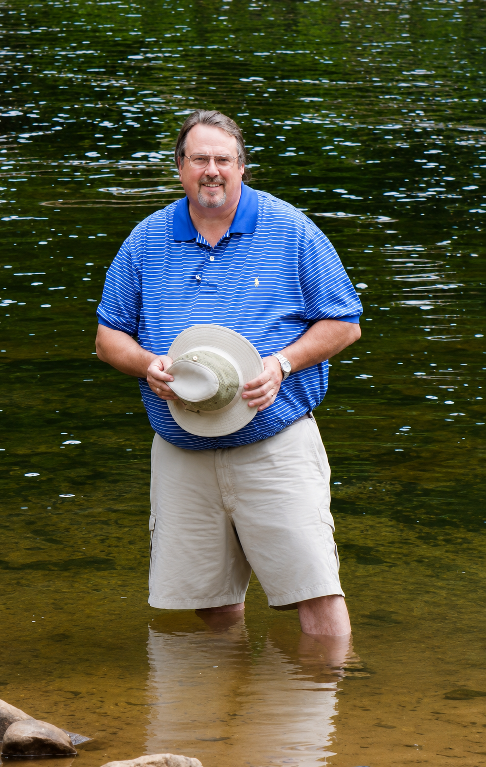 Man in blue striped polo shirt and beige shorts standing in shallow water, holding a wide-brimmed hat, with a calm lake in the background.
