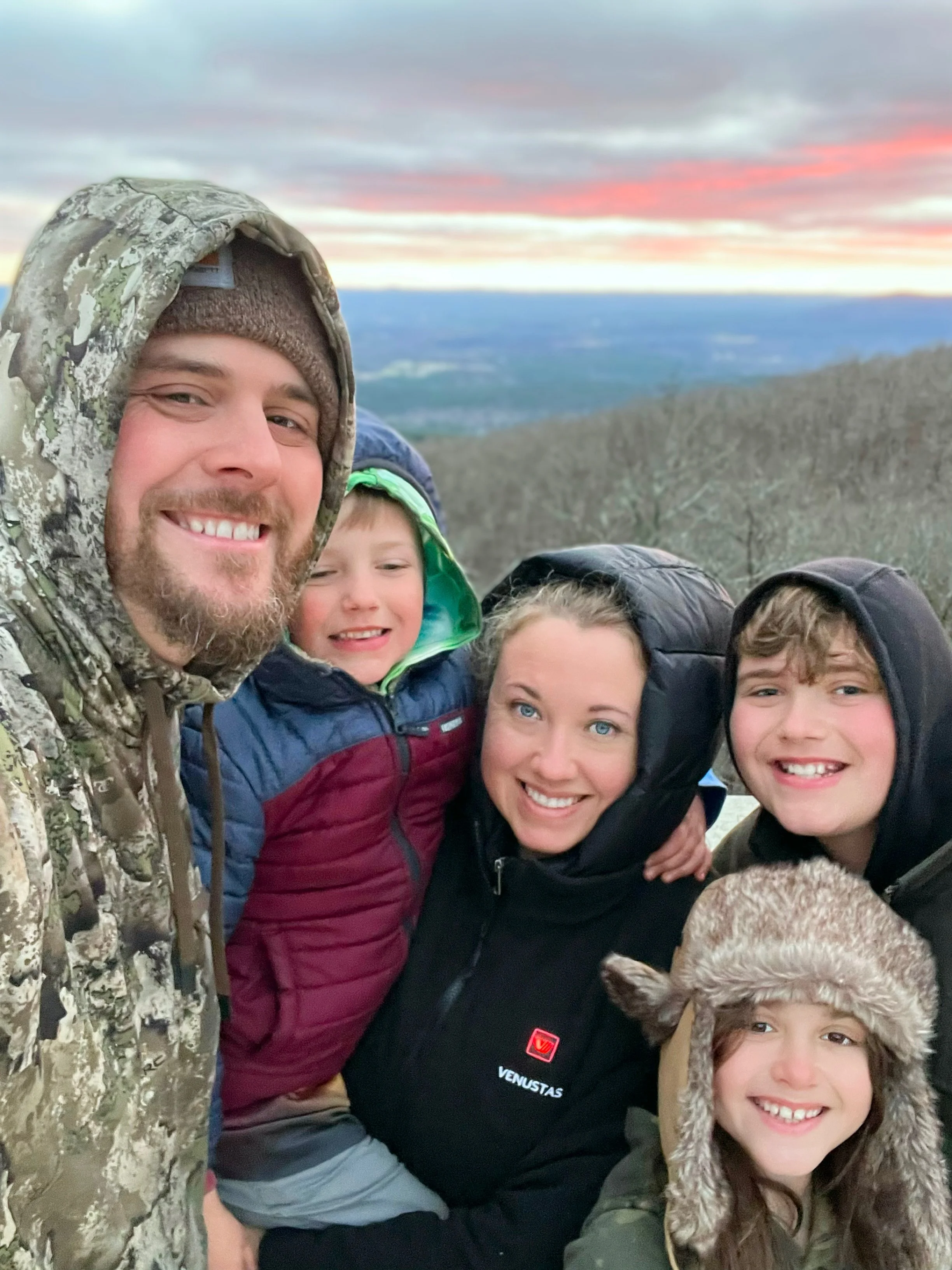 A group of smiling people, including children and adults, outdoors during sunset or sunrise. They are dressed warmly in jackets, hats, and furry accessories. The landscape behind them features a scenic view of rolling hills and a colorful sky with clouds.