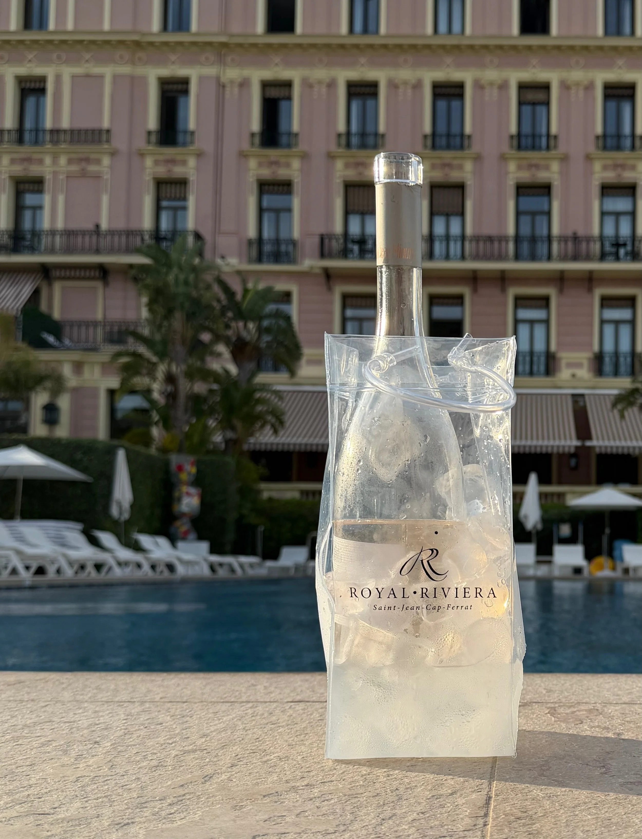 A clear plastic bag with an ice-cold bottled beverage labeled 'Royal Riviera' in front of a swimming pool, with a pink building with multiple windows and balconies in the background.