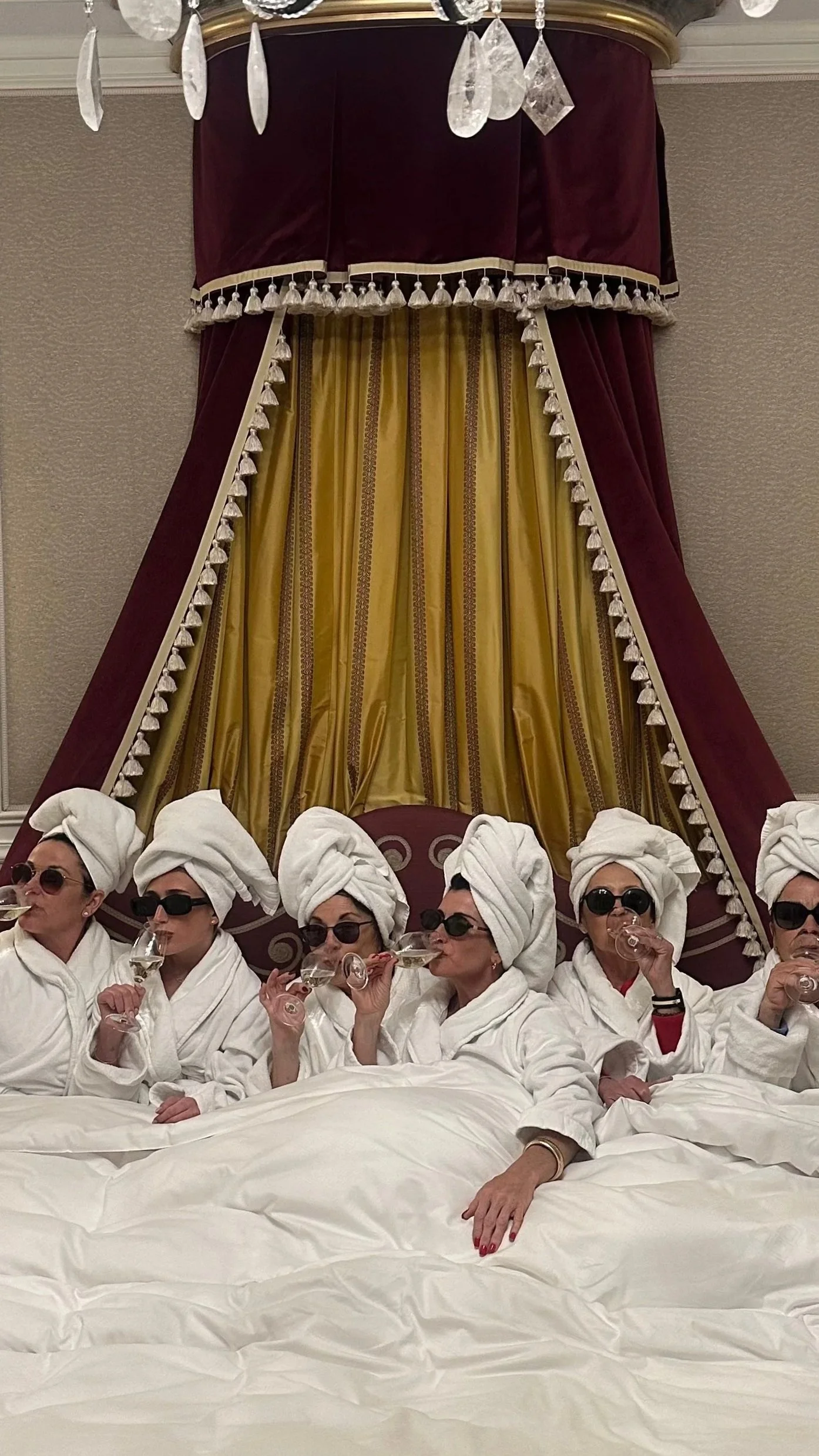 A group of women dressed in white bathrobes and white towels wrapped around their heads, wearing dark sunglasses, sitting at a spa or hotel room, drinking champagne, with a luxurious bed and an ornate canopy with gold and maroon curtains in the background.