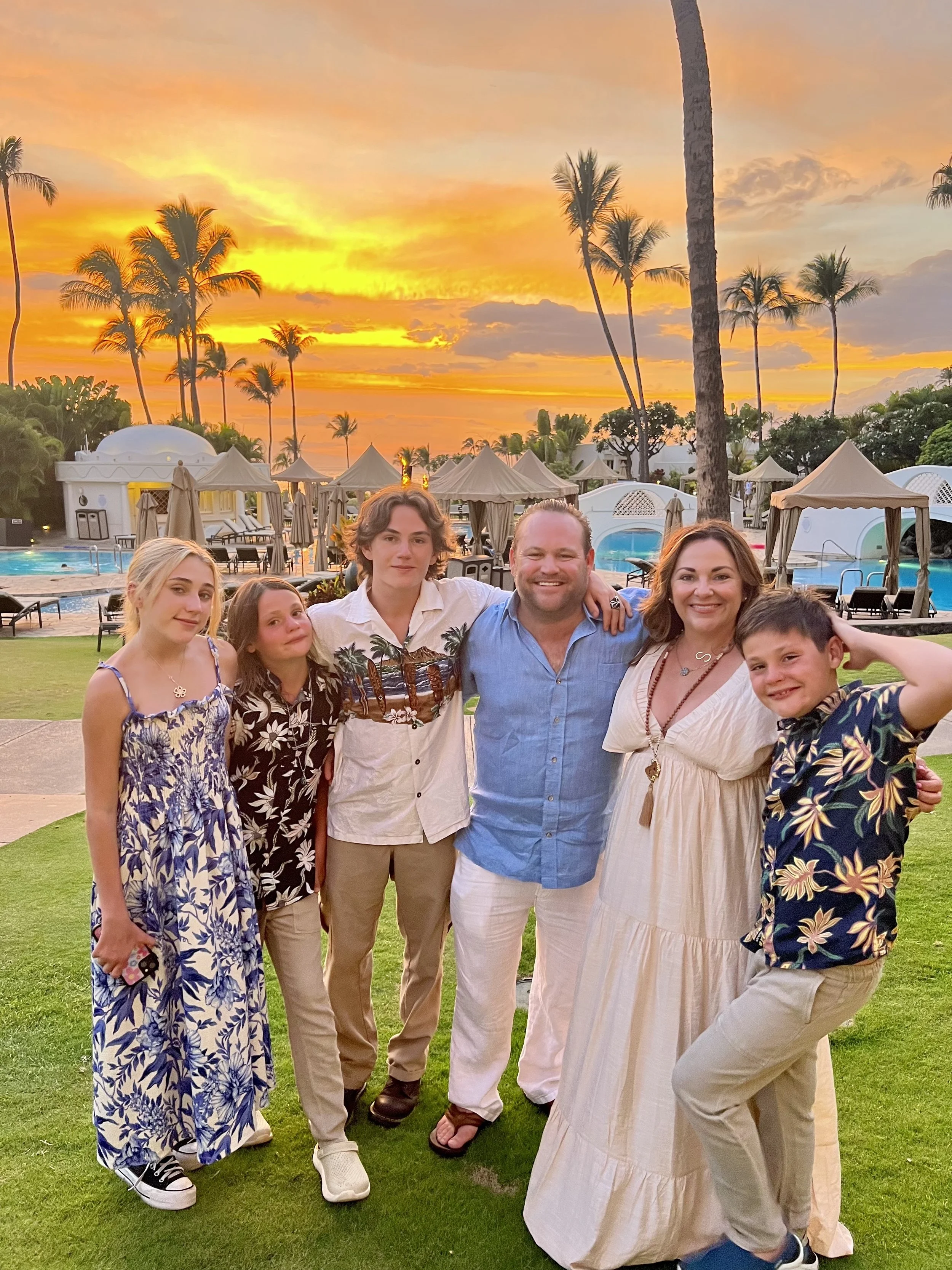 Group of seven people posing together outdoors at sunset, with palm trees and cabanas in the background, on a lawn next to a pool.
