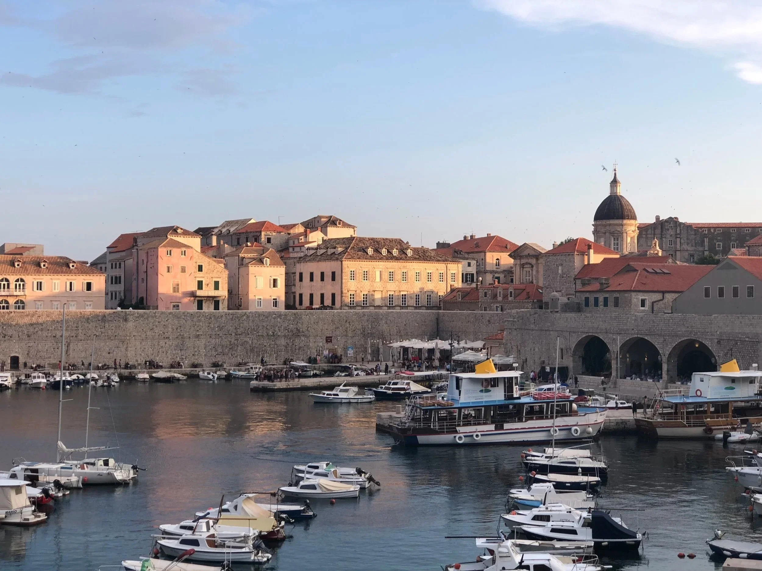 A harbor with multiple boats docked in the water. In the background, there are historic buildings with red-tile roofs and a church tower with a dome and bell tower. The sky is partly cloudy.