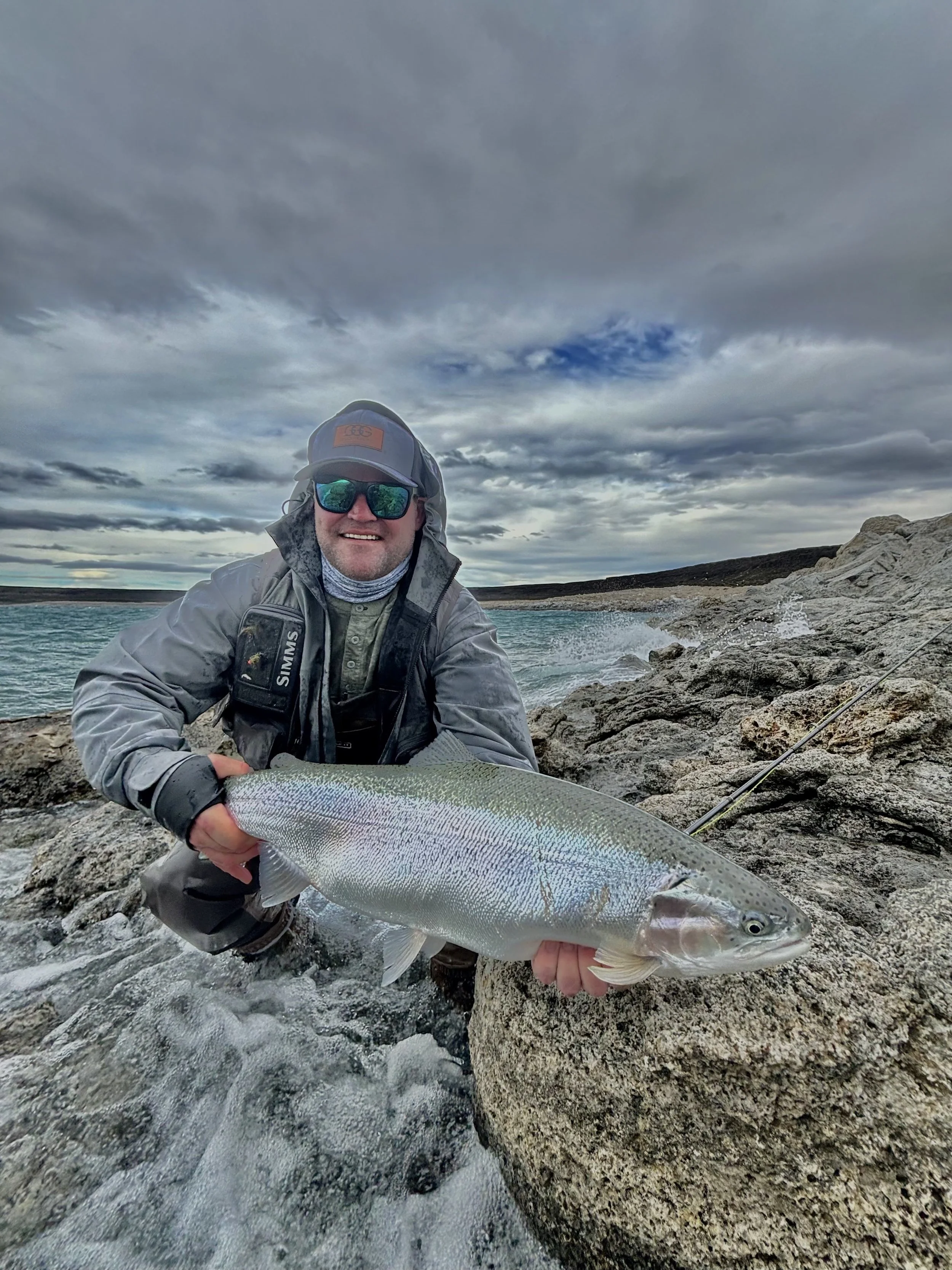 A man wearing sunglasses, a hat, and outdoor fishing gear is kneeling on rocky shore, holding a large fish. The background shows a cloudy sky and a body of water with waves crashing against rocks.