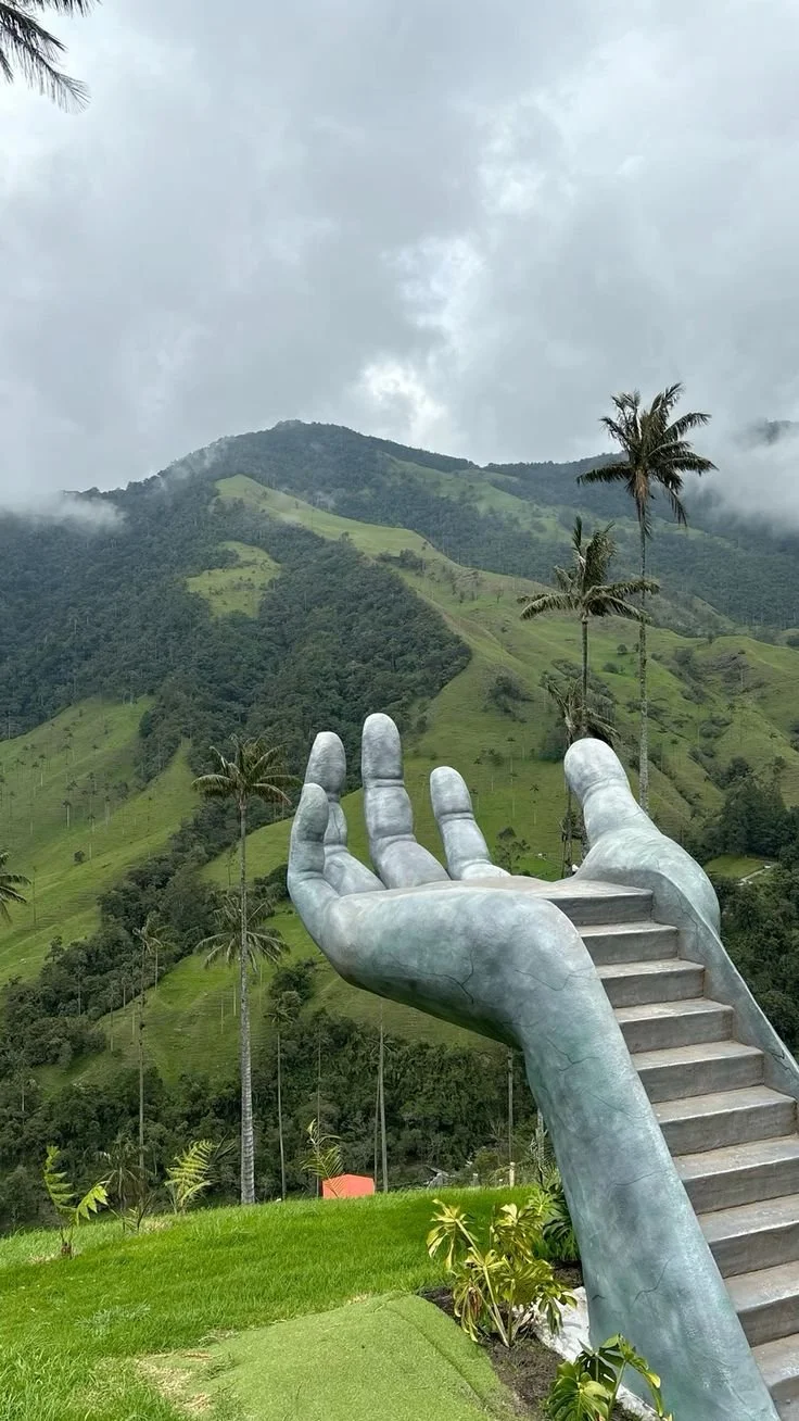Escultura de una mano gigante que tiene escaleras en la palma, con fondo de montañas verdes y árboles de palma. Cielo nublado.