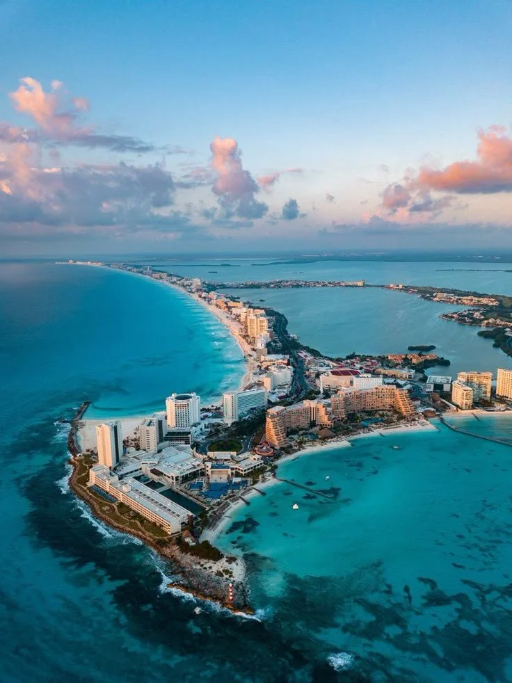 Vista aérea de un destino turístico en la playa, con altos edificios, arena blanca y agua azul turquesa, bajo un cielo con algunas nubes.