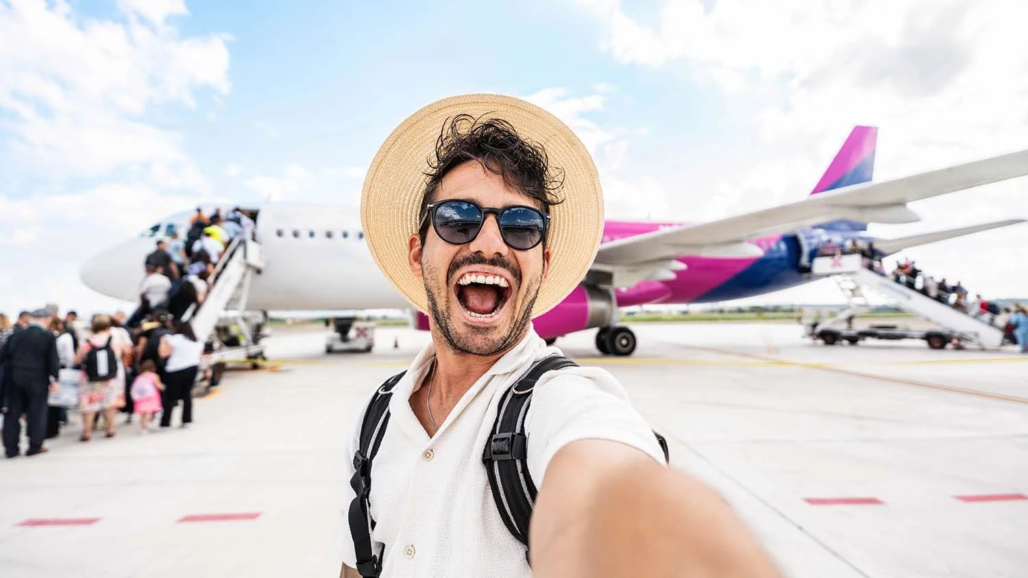 Hombre sonriente tomando selfie en aeropuerto con avión y pasajeros subiendo a bordo en el fondo.