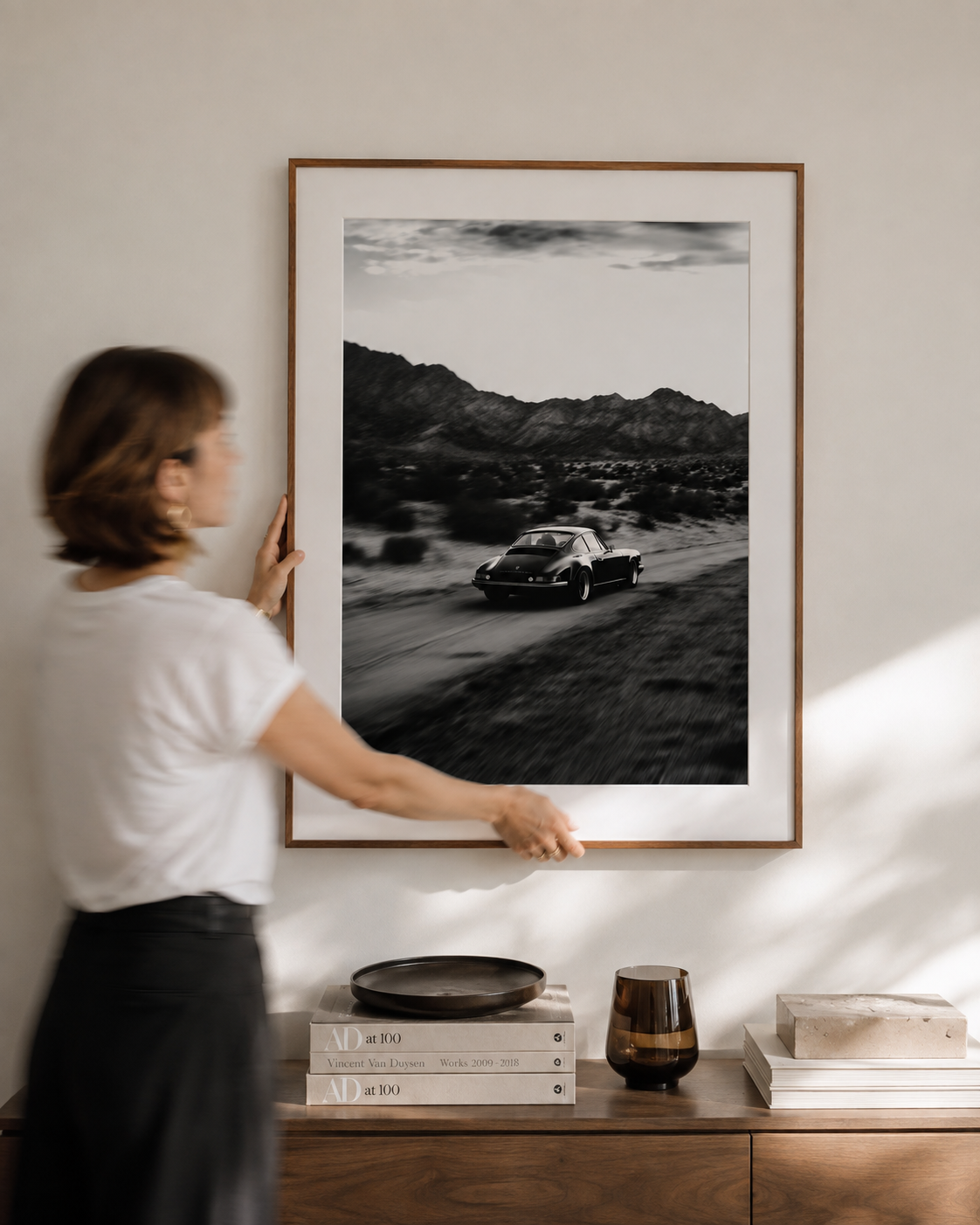A motion-blurred woman carefully hangs a large framed black-and-white artwork in a bright modern interior, styled with a walnut console, design books, and minimalist decorative objects.