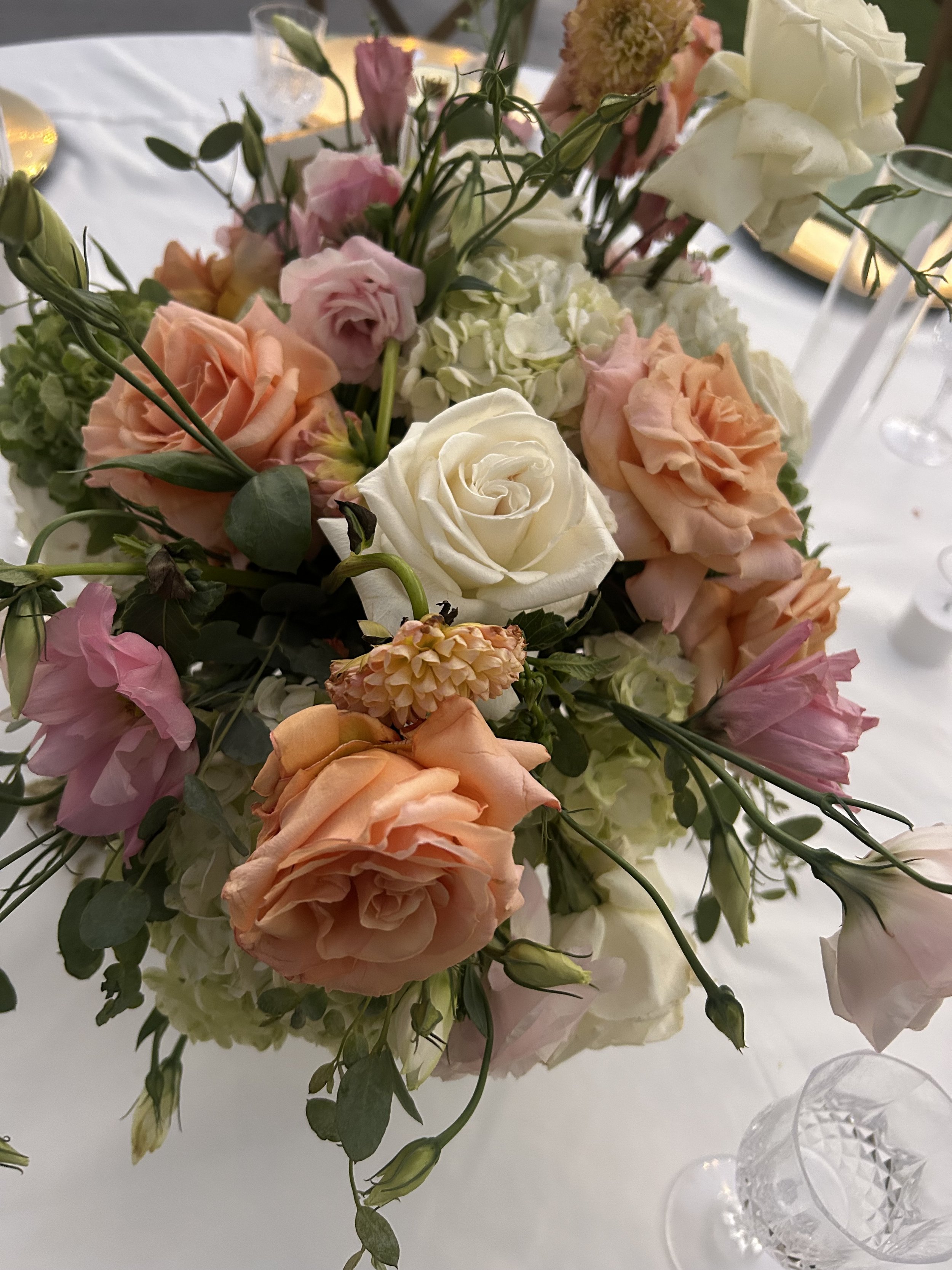 A floral centerpiece with pink, peach, white roses, and green foliage on a white tablecloth, surrounded by glassware and candles.