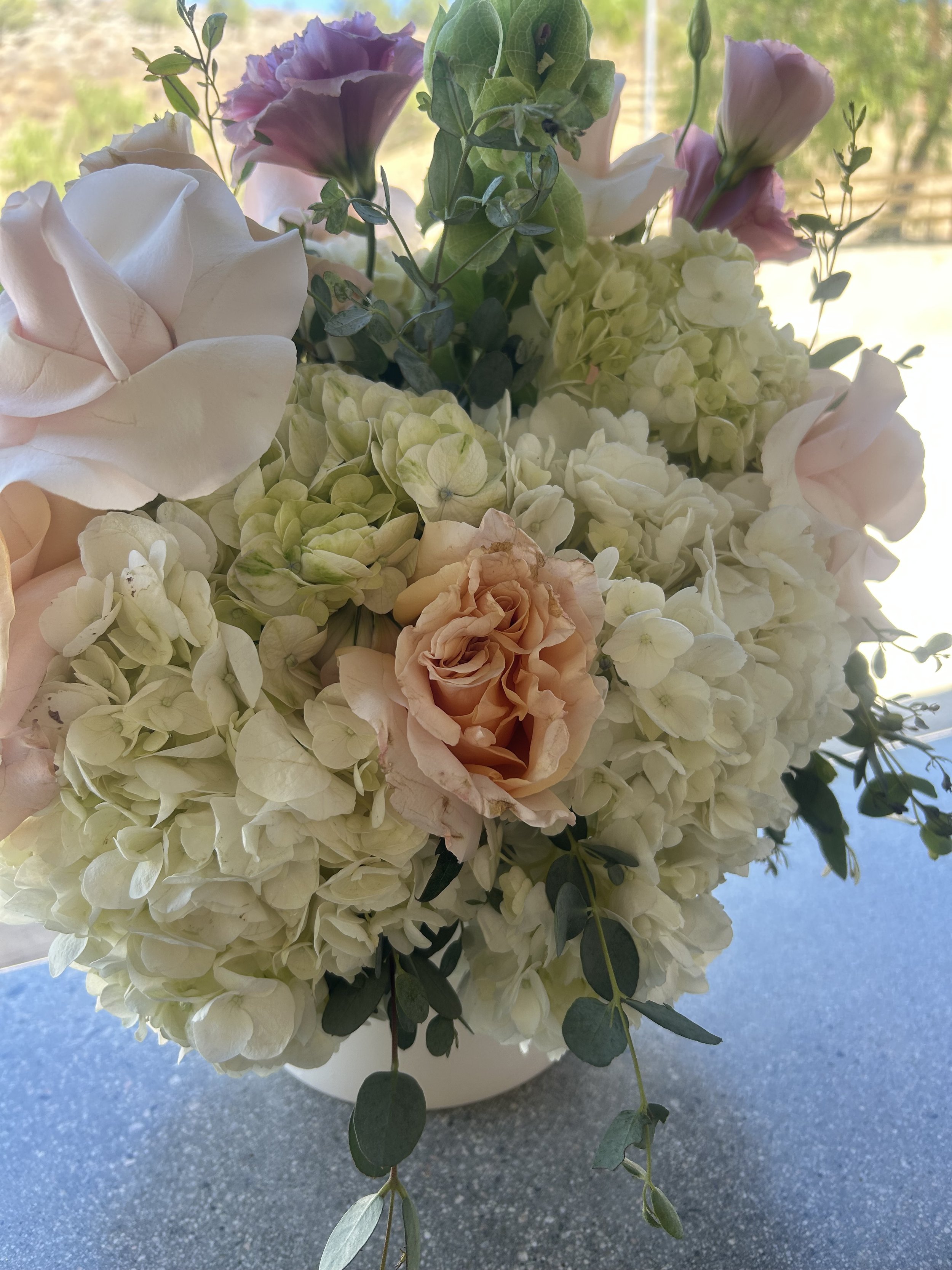 A bouquet of various flowers including white hydrangeas, blush pink roses, and purple lisianthus, with greenery, placed in a white vase on a speckled countertop.