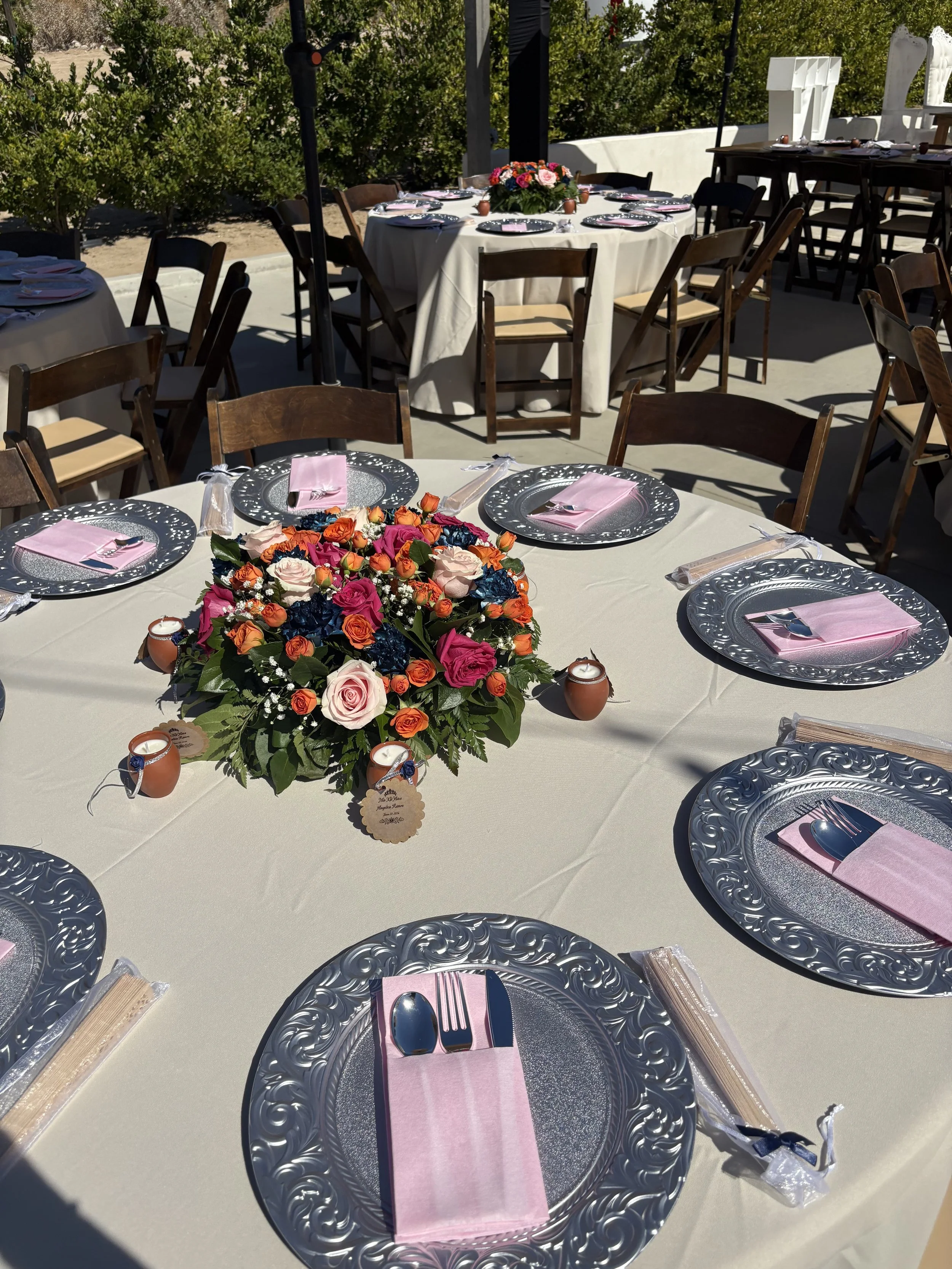 Round banquet table set for a formal event outdoors, decorated with a large centerpiece of colorful roses, pink napkins, silverware, and ornate silver plates, with multiple wooden chairs around it.