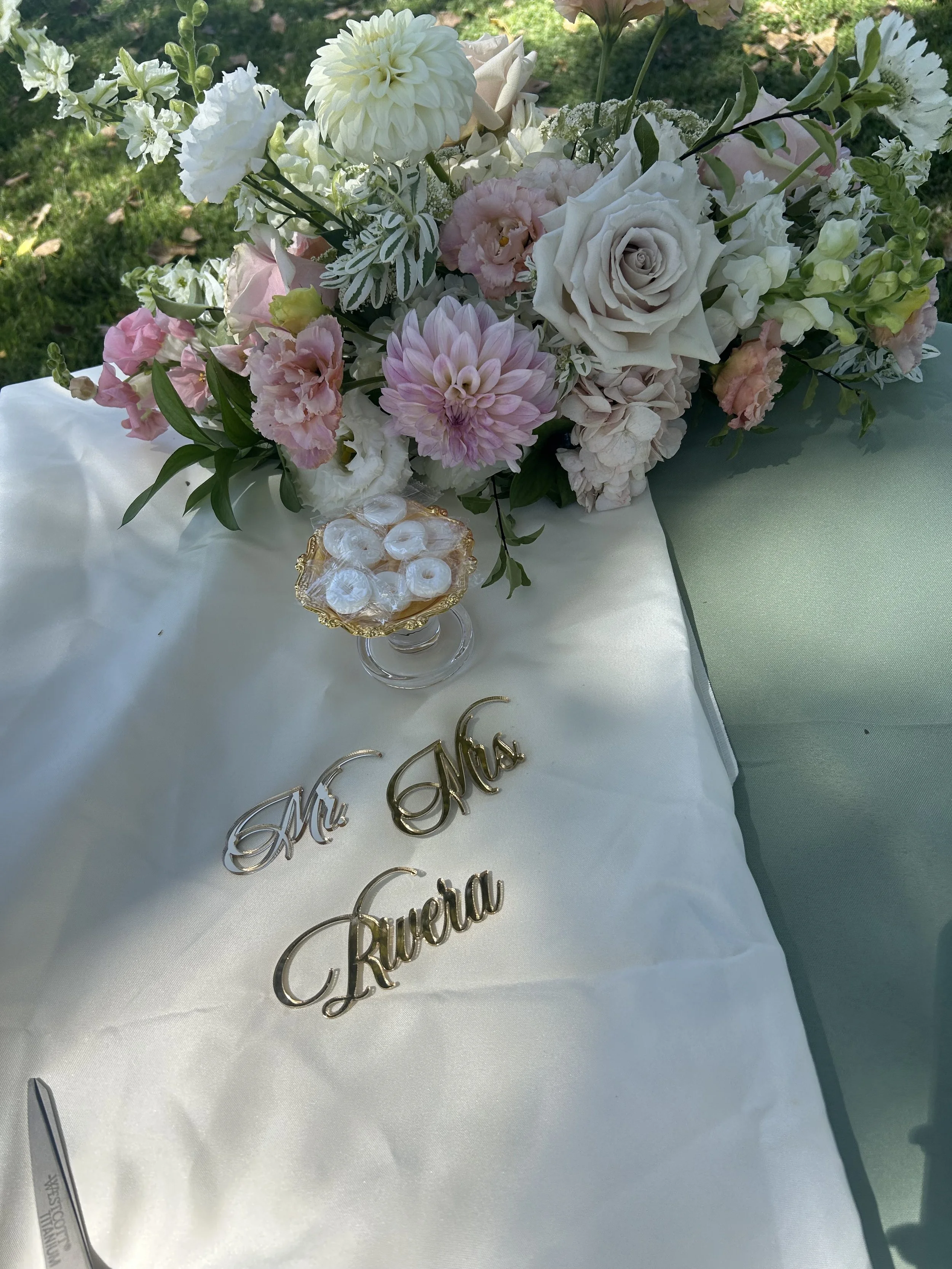 A floral arrangement of white, pink, and cream-colored flowers on a table with a white tablecloth, alongside gold and silver decorative words and jewelry, outside in a garden setting.