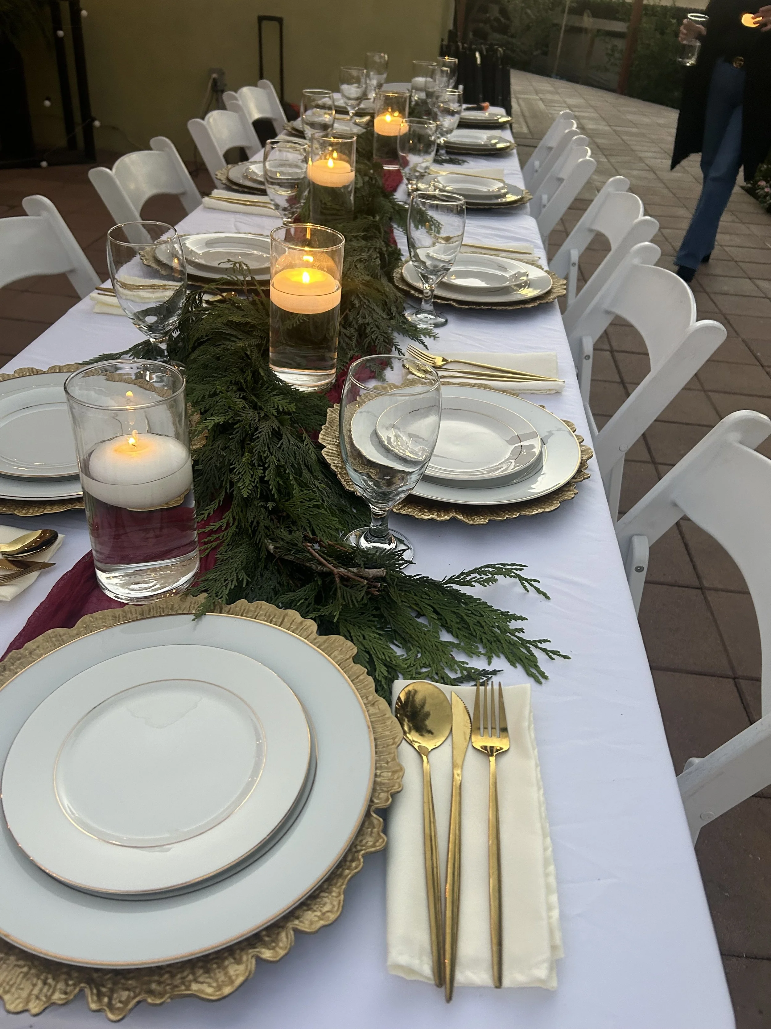 A long rectangular dining table set for a meal with white plates with gold rims, gold cutlery, and white napkins. The table has a white tablecloth and a green garland with candles as a centerpiece. There are water glasses at each place setting and some candles lit. The setting appears festive, possibly for a holiday gathering.