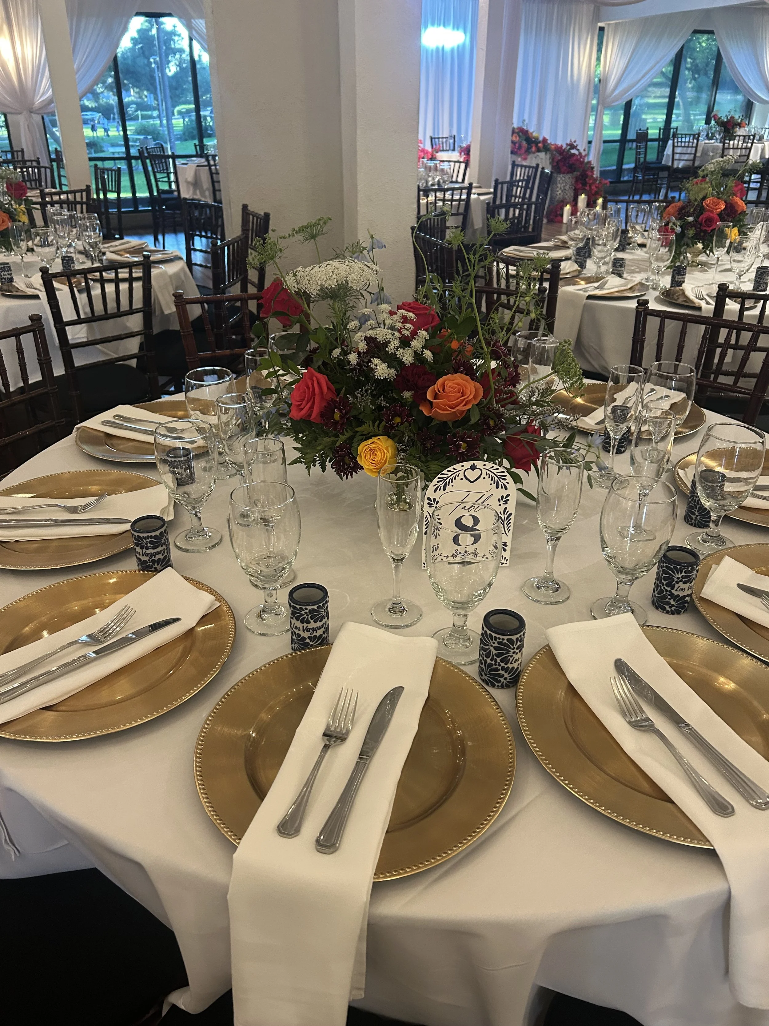 Elegant banquet table setup with white tablecloth, gold chargers, silverware, white napkins, and floral centerpieces, inside a decorated event hall with large windows and draped curtains.