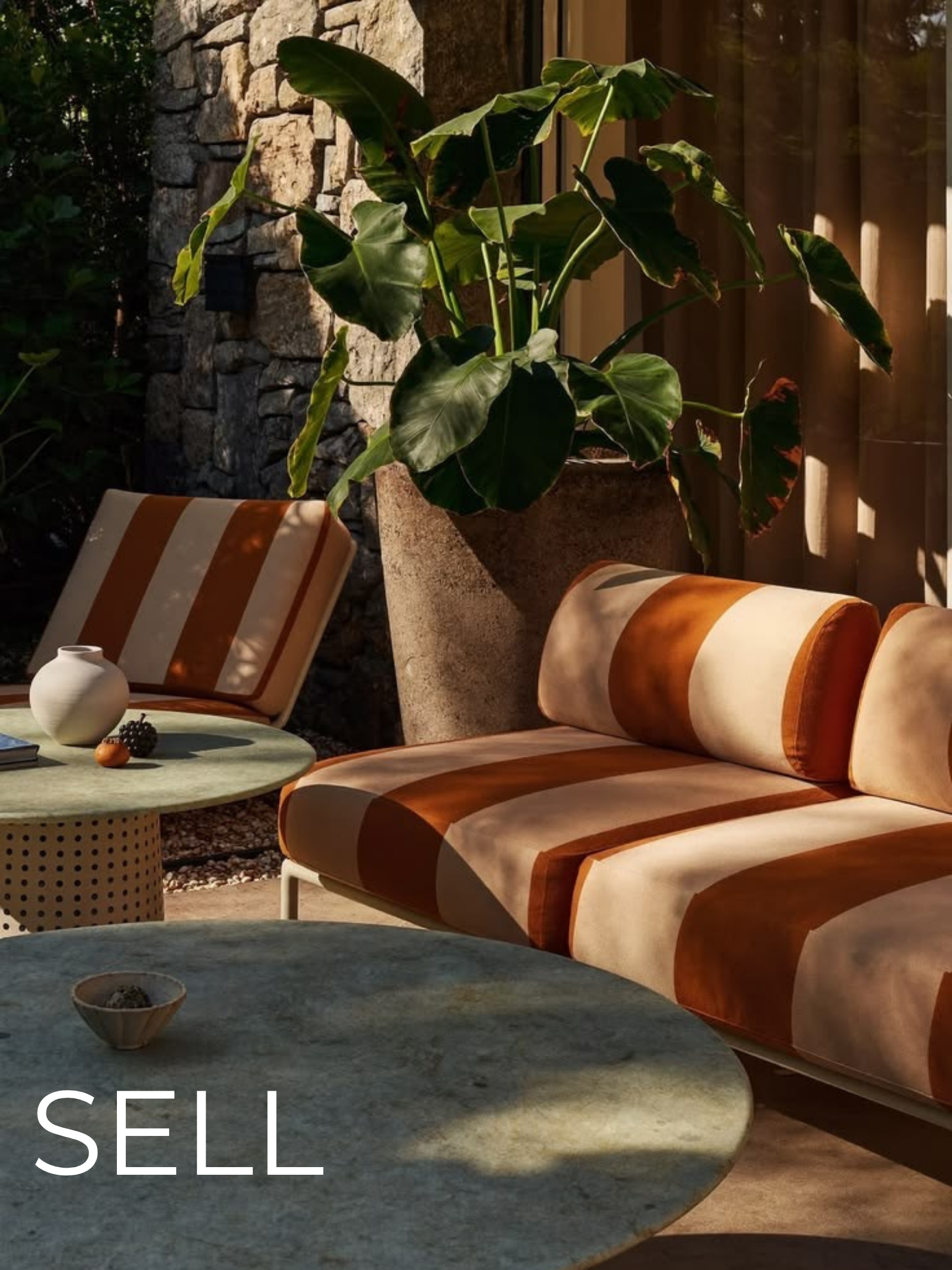 A cozy outdoor seating area featuring a striped sofa with orange and white cushions, a green round table with a small bowl and black berries, and a large green leafy plant in a pot, illuminated by warm sunlight.